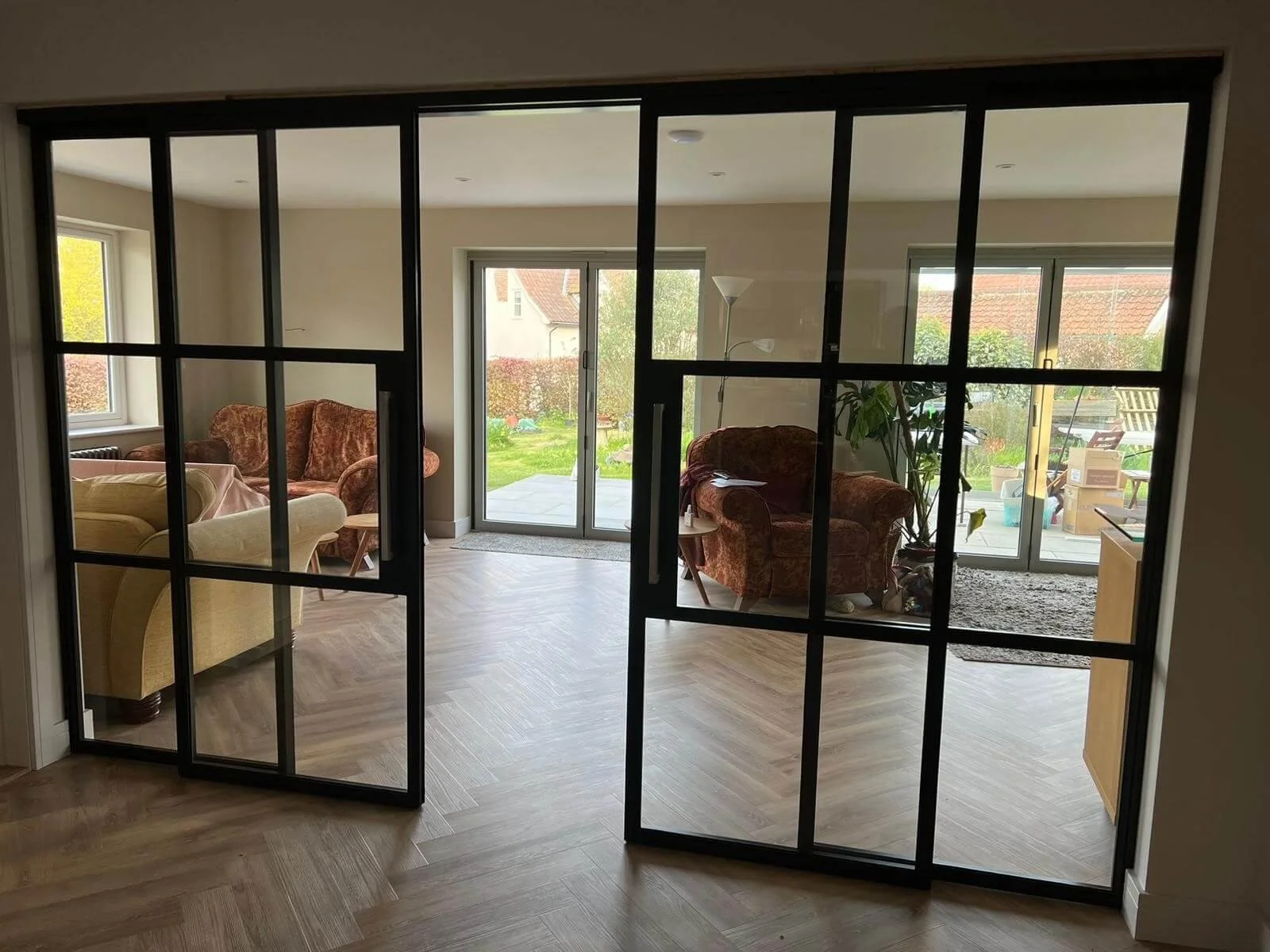 View into a living room seen through black-framed glass folding doors. The room has beige walls, hardwood flooring, several armchairs, a large window, sliding glass door, a tall floor lamp, and a potted plant. Outside the sliding door, there is a patio with outdoor furniture, and a garden with shrubs and trees.