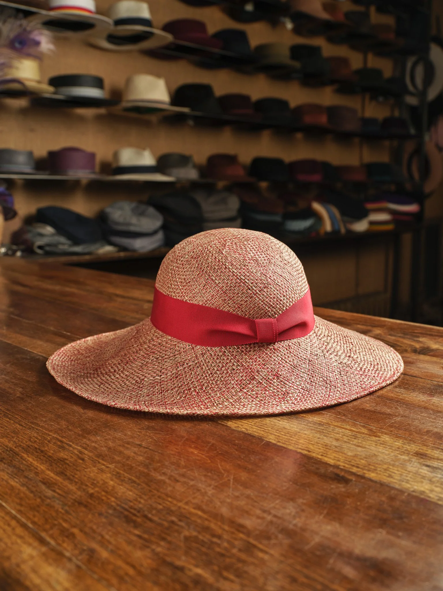 A pink and beige wide-brimmed straw hat with a pink ribbon, resting on a wooden table in a hat store. Shelves in the background display various hats in different colors and styles.
