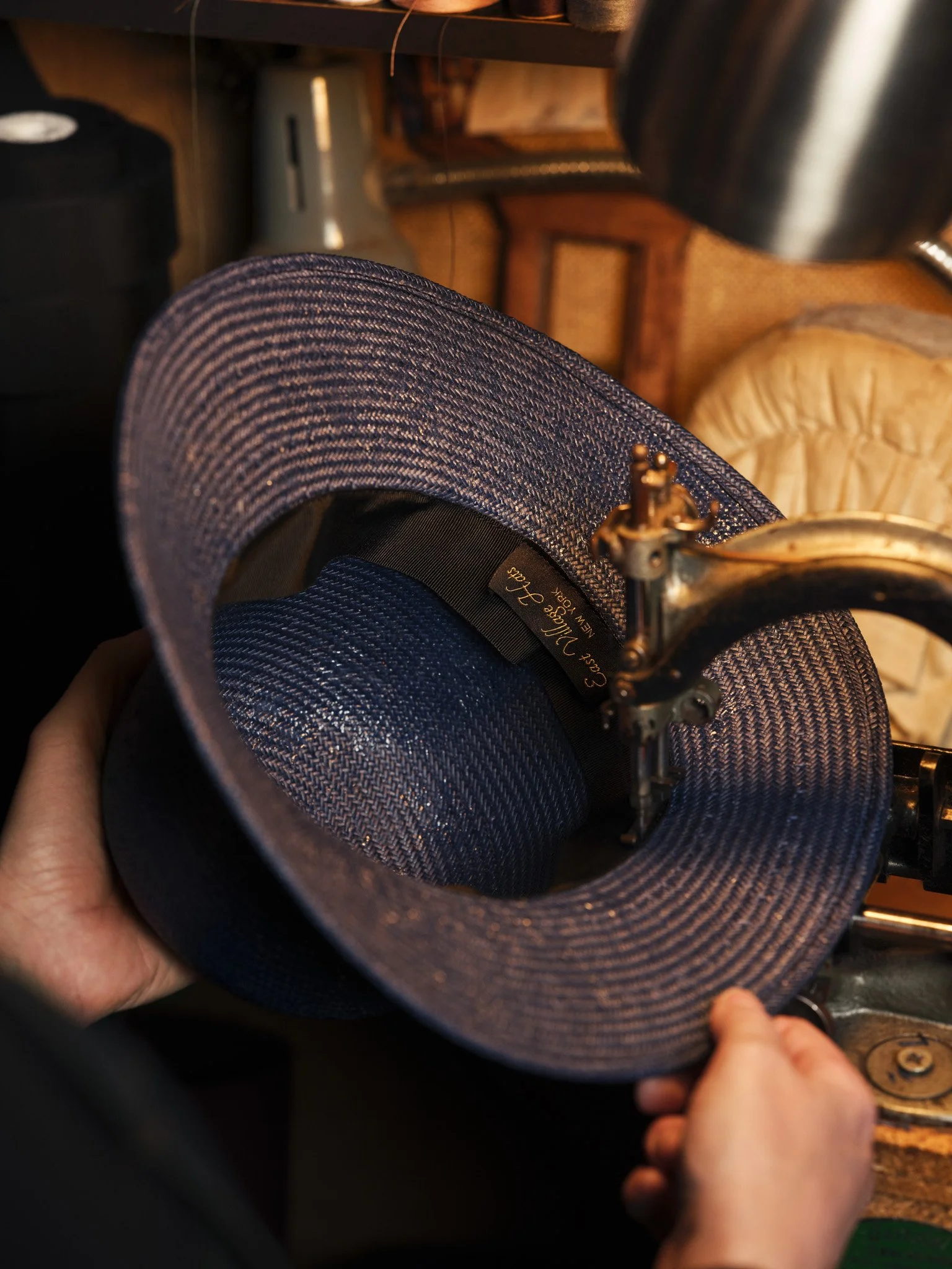 Close-up of a person sewing or repairing a wide-brimmed hat with a pin and sewing machine, with a wooden surface and a coffee mug in the background.