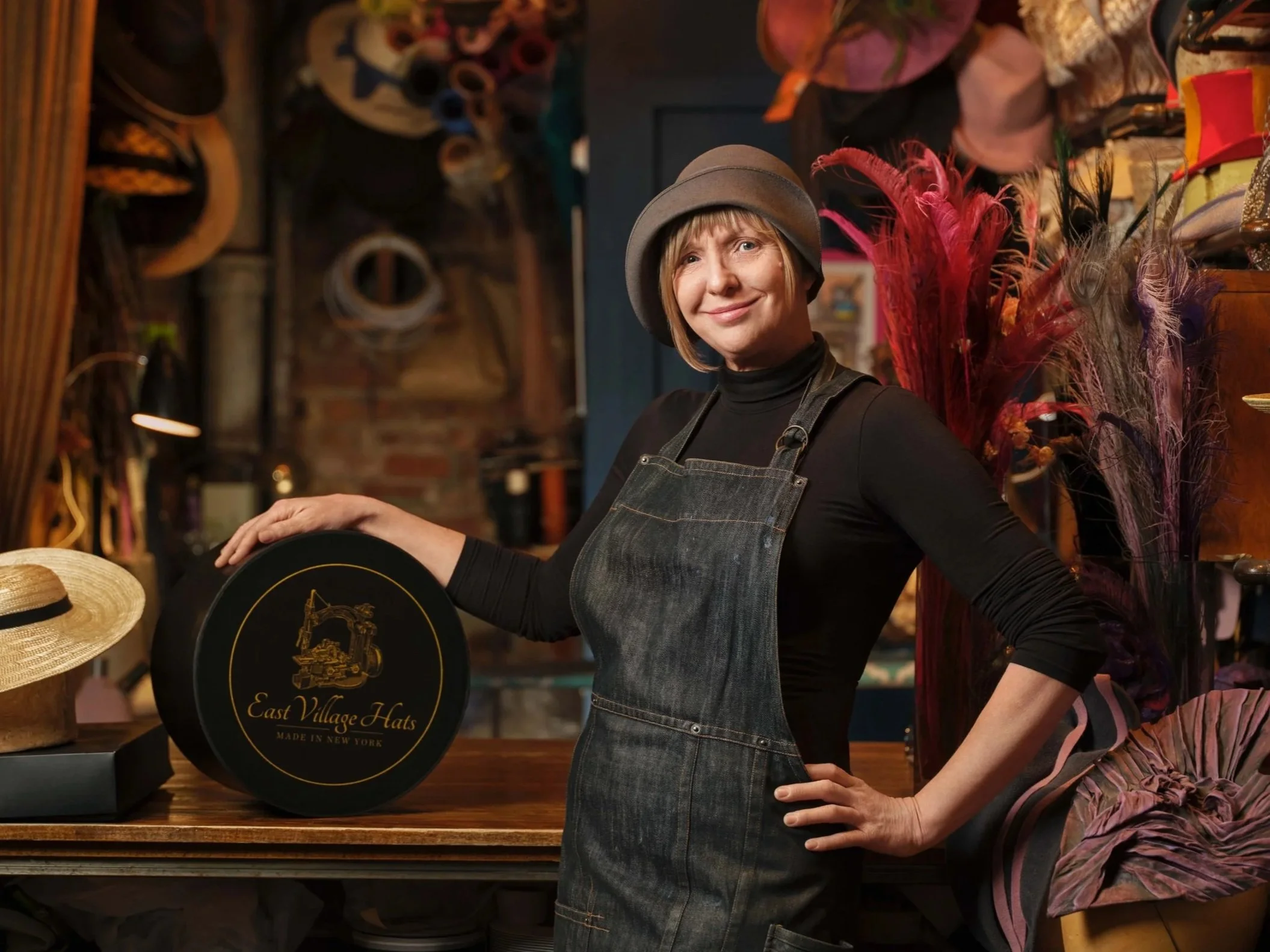 A woman with short hair wearing a brown hat, black turtleneck, and apron, smiling in a hat shop surrounded by hats and colorful feathers.
