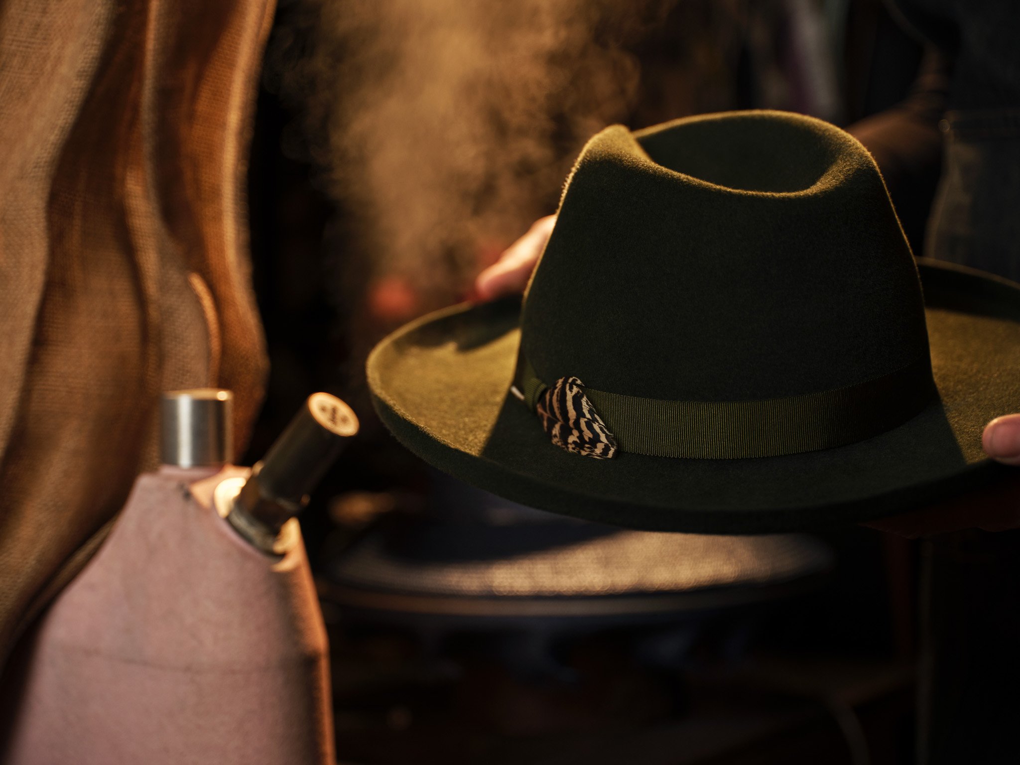 A black fedora hat with a decorative band, resting on a surface surrounded by a brown bag and other objects, with a person blurred in the background.