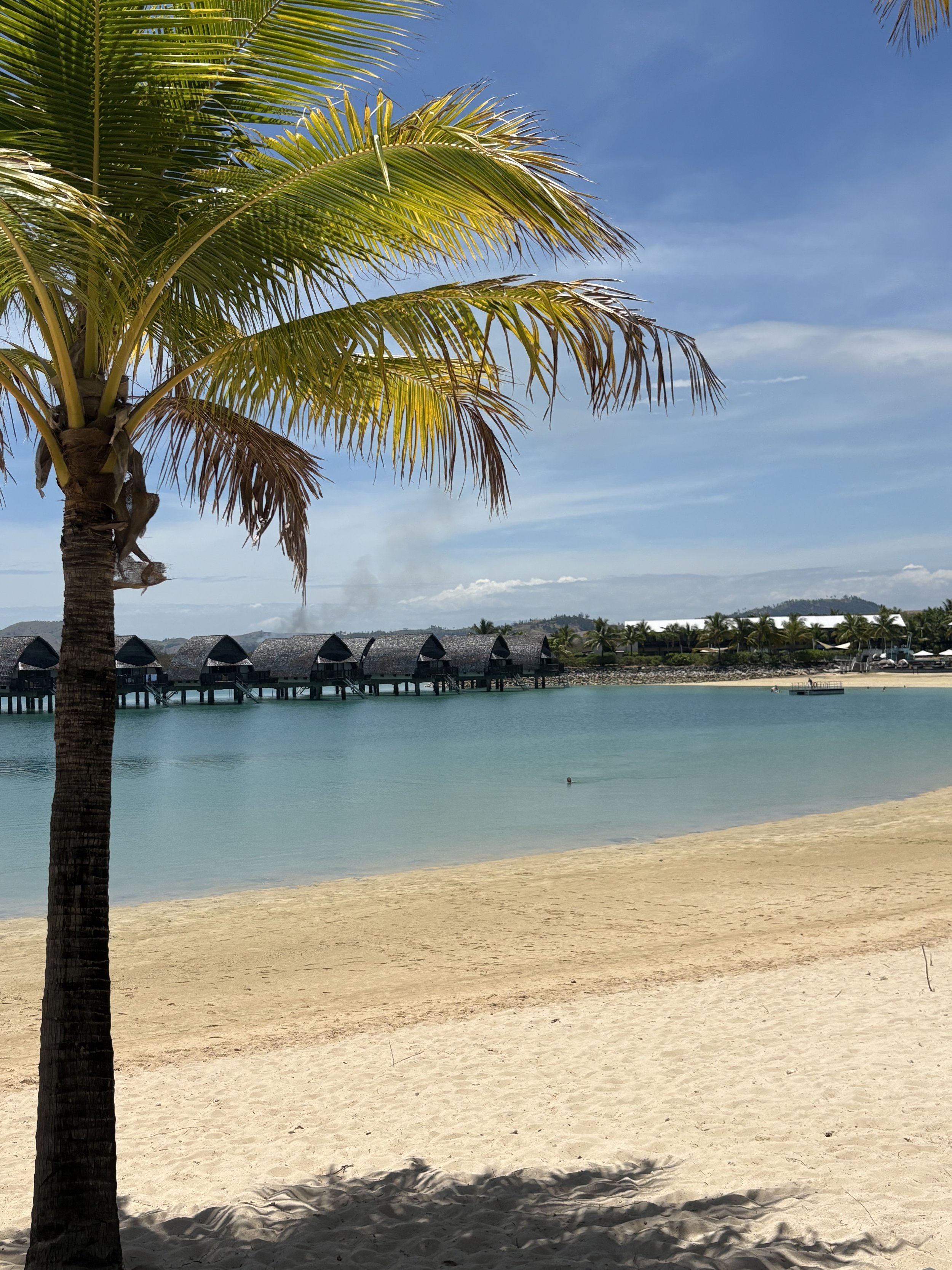 A tropical beach scene with a palm tree in the foreground, over a sandy beach, clear water, and overwater bungalows in the distance under a partly cloudy sky. Marriott Mami Bay resort in Fiji.