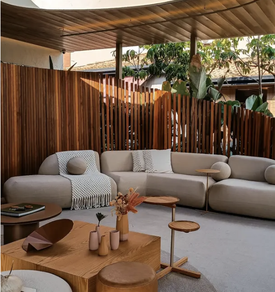 Living room with a modern off-white sectional sofa, a wooden coffee table with decorative vases, and a wooden partition wall. There are trees visible outside through an open ceiling with wood paneling.