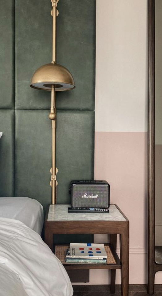 A bedside table with a Marshall speaker and books, a bed with white sheets, a green padded wall with a gold wall lamp, and a partial view of a mirror. Luxury hotel accommodations.