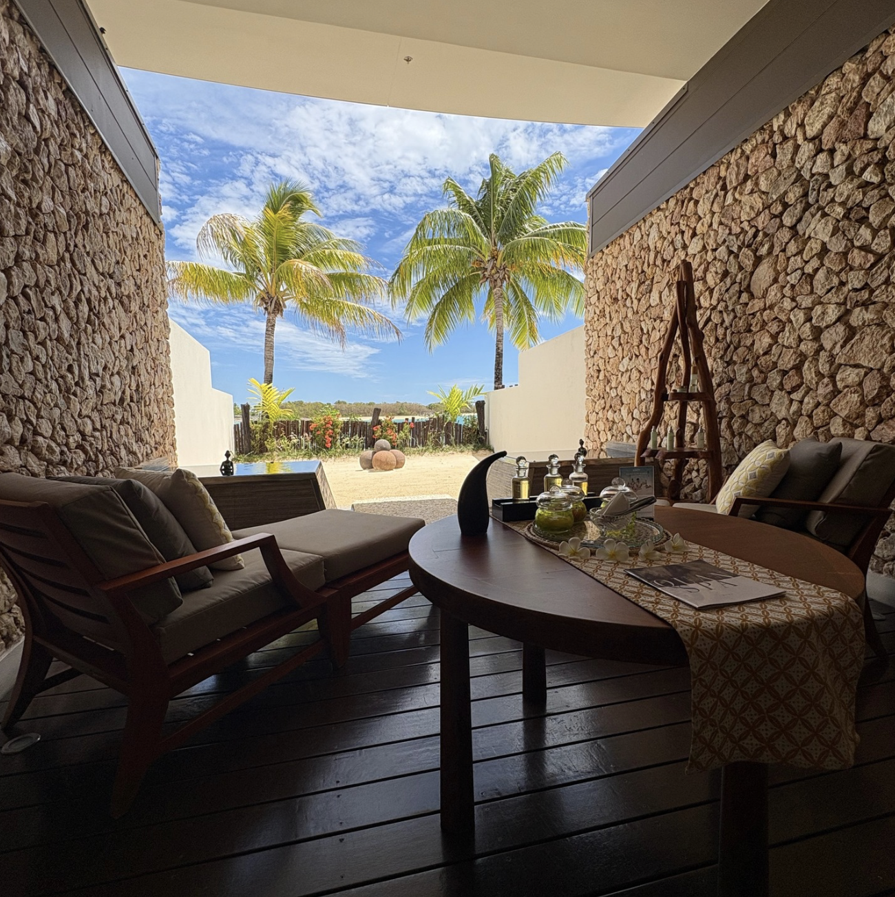 View of a tropical outdoor scene with palm trees, sandy ground, and blue sky, seen from a shaded patio with wooden floor, outdoor furniture, and a decorative table setting.  Spa registration area at Mami Bay Resort in Fiji.