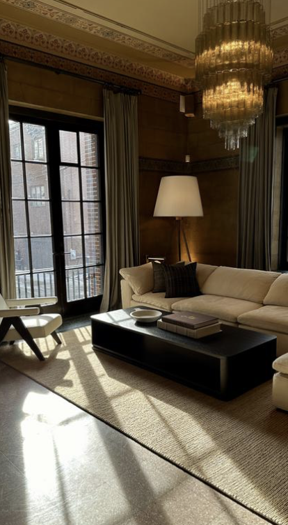 Living room with large windows, a cream sofa with black pillows, a black coffee table with books and a bowl, a white floor lamp, a chandelier, and curtains.  Corporate hotel travel.