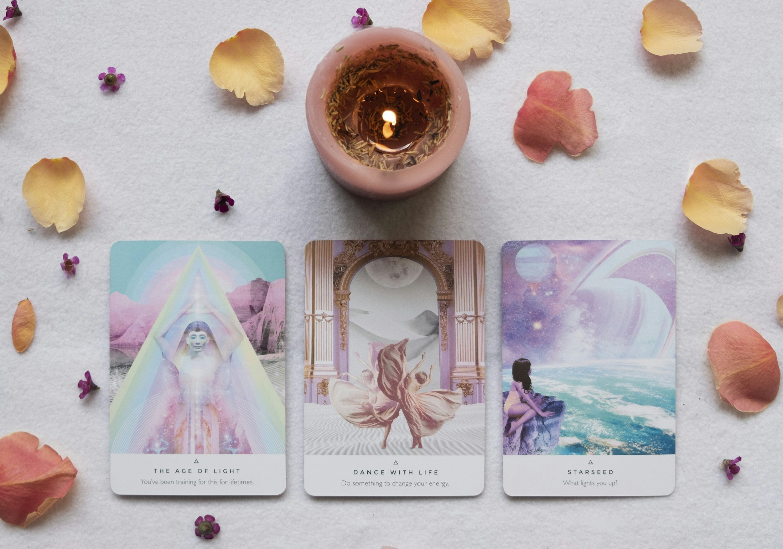 Three tarot cards laid out on a white surface with rose petals and small purple flowers scattered around. The first card depicts a light figure with a rainbow and mountainous background titled 'The Age of Light.' The second card shows a woman with flowing hair and a horse with a desert background titled 'Dance with Life.' The third card features a cosmic scene with a silhouette of a person against an ocean background titled 'Starseed'.