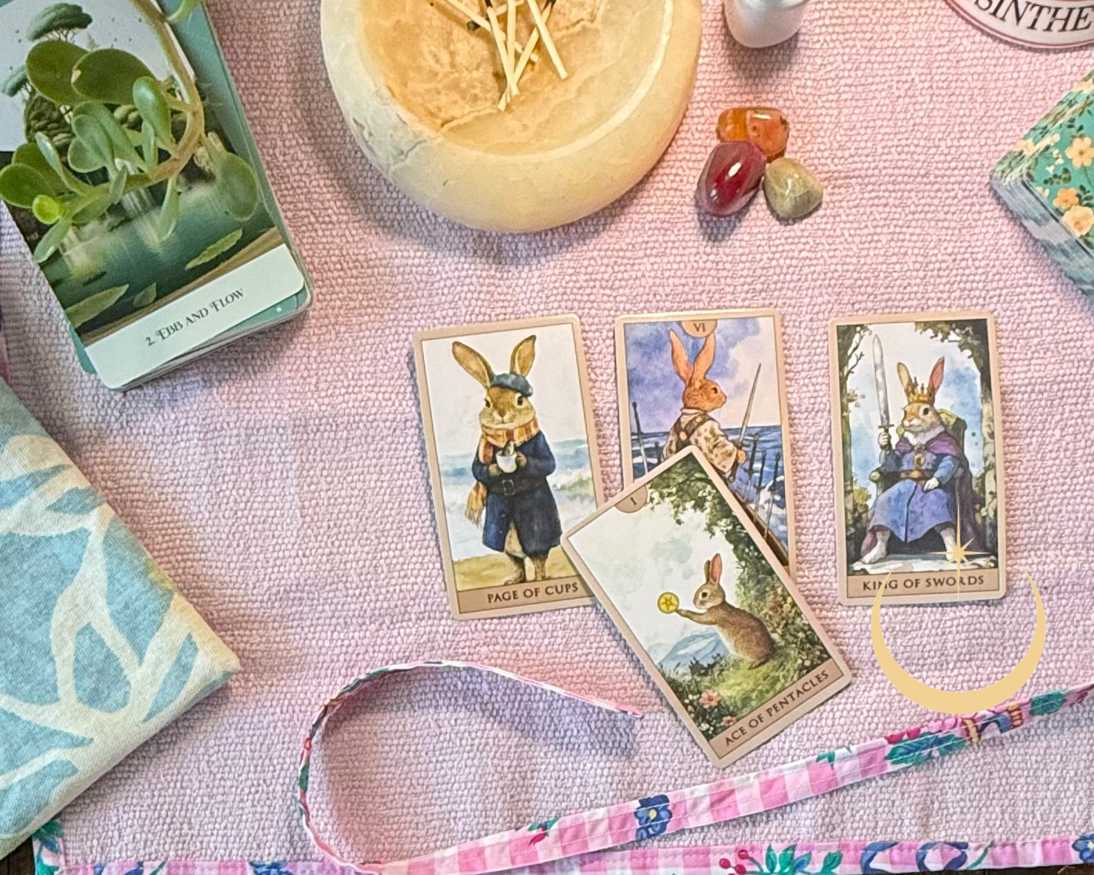 Tarot cards featuring rabbits in royal and adventurer roles, lying on a pink textured cloth with a potted plant, a candle holder, and decorative stones nearby.