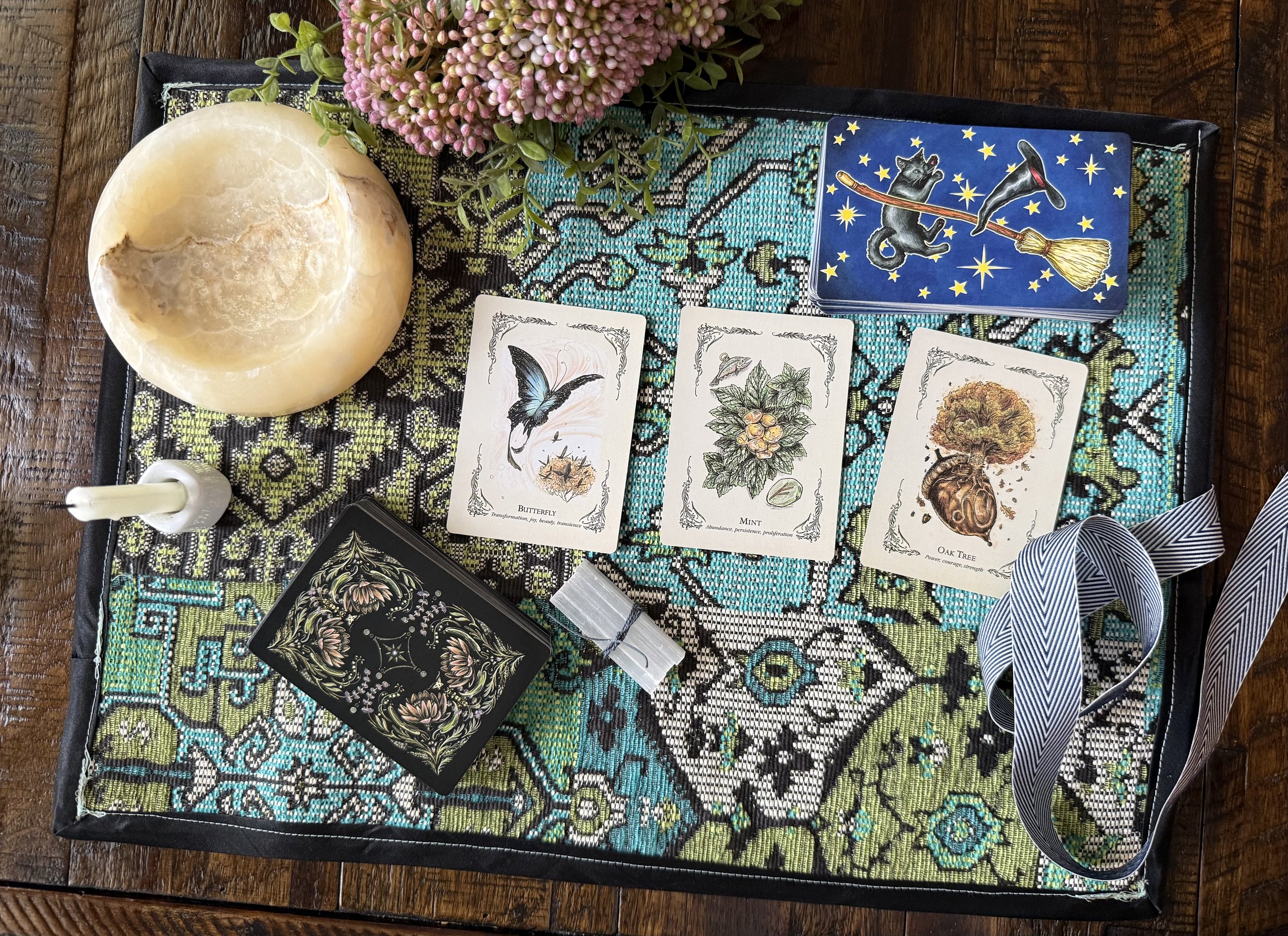 A lunar light lamp, a bundle of pink flowers, tarot cards featuring a butterfly, mint, and oak tree, a black book with floral design, a white candle, and a black and white striped ribbon are arranged on a decorative fabric tray on a wooden surface.