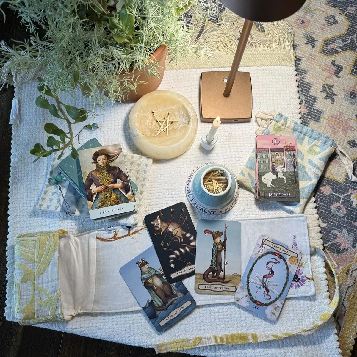 A tarot reading setup on a white cloth featuring tarot cards, candles, a flower-patterned cloth pouch, a jar with matches, a potted plant, a small clock, and a lamp with a beige base.