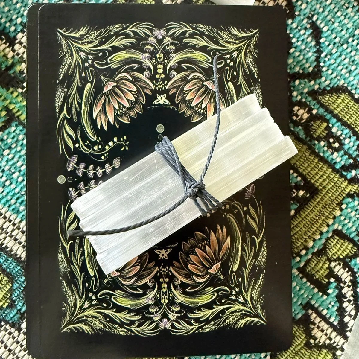 A closed notebook with ornate golden floral patterns on a black cover, placed on colorful woven fabric. A bundle of white sticks, tied with a gray string, rests on the notebook.