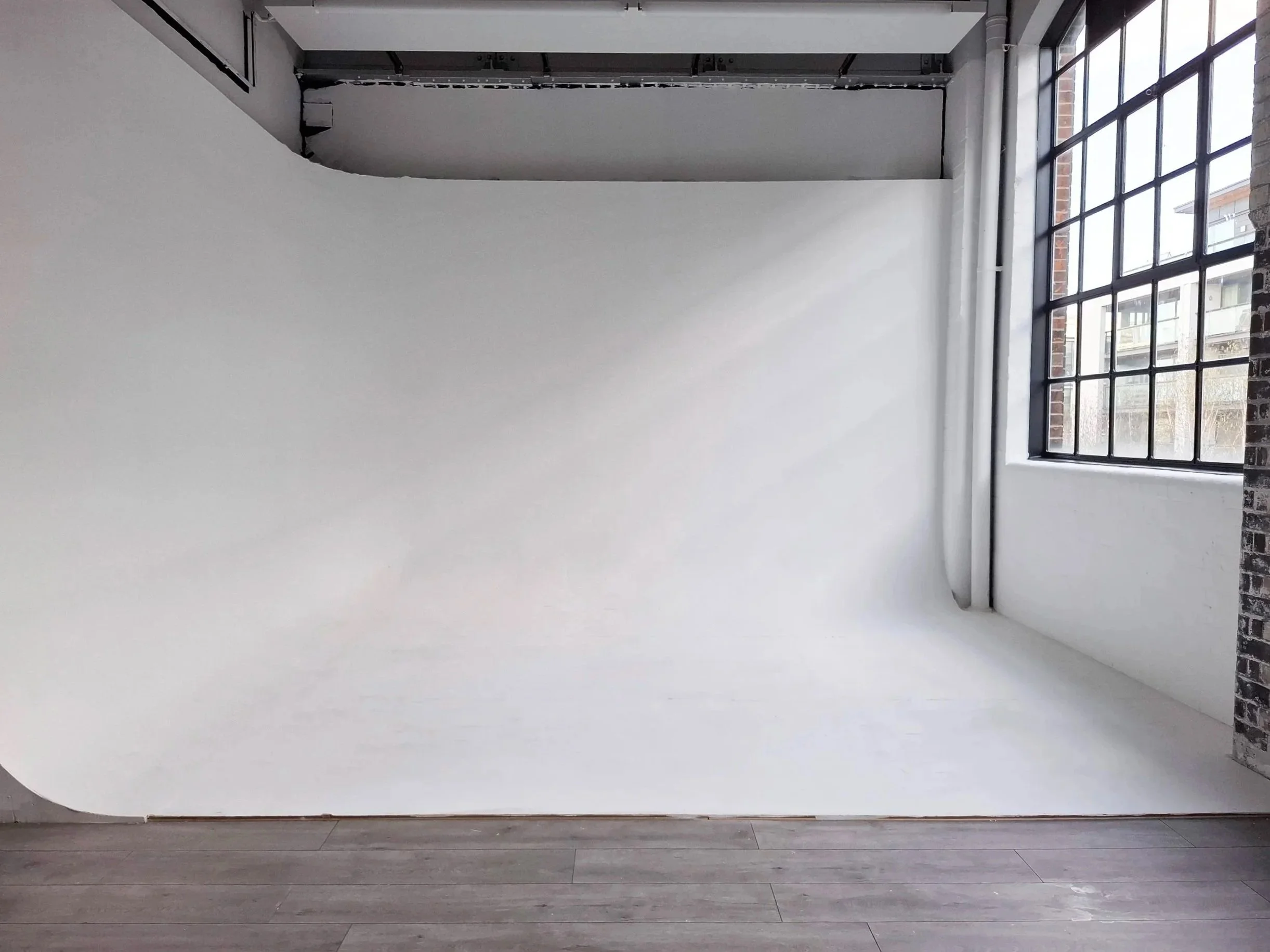 An empty photo studio with a white backdrop, large grid window on the right allowing natural light, and hardwood flooring.