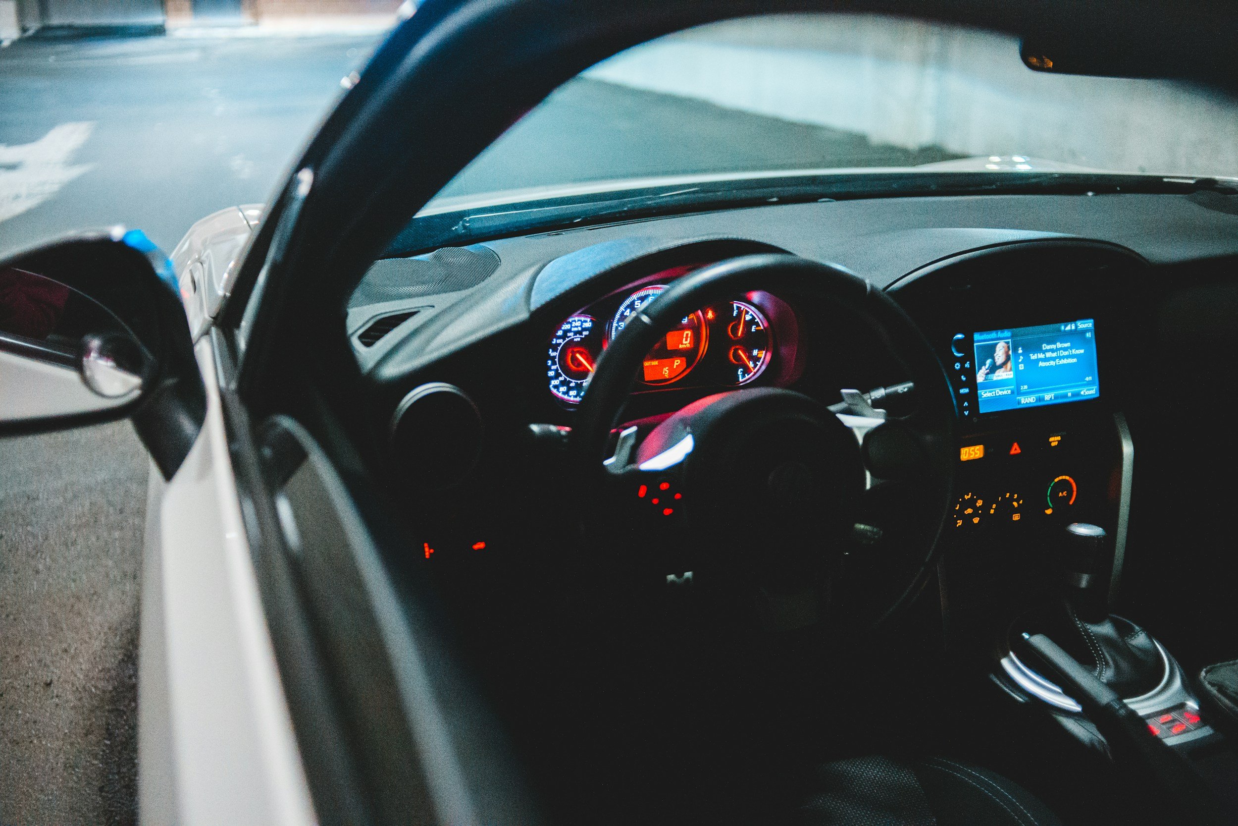 View of a car's dashboard through the windshield, showing speedometer, fuel gauge, and infotainment screen at night, with the street outside faintly visible.
