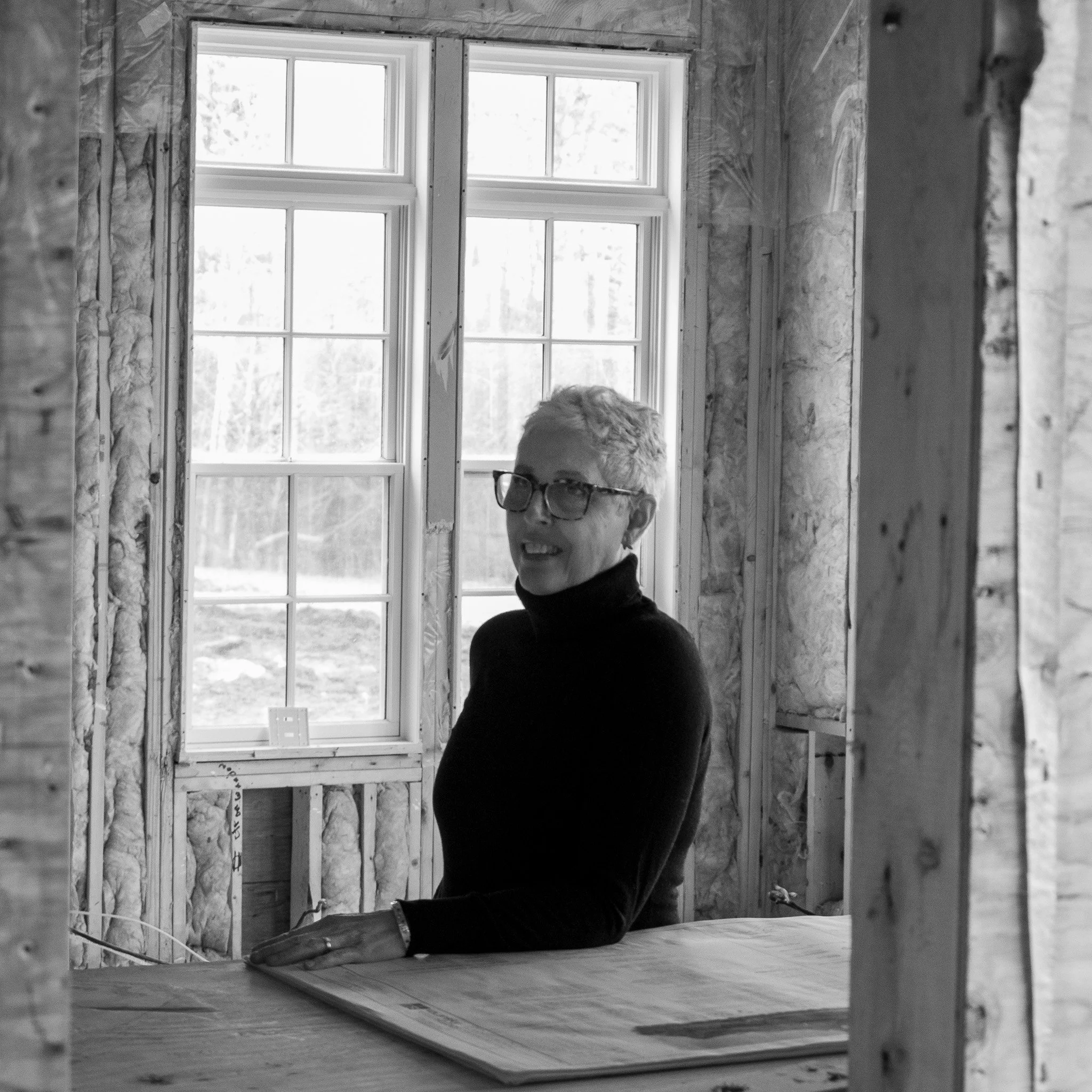 Black and white photo of a woman with short hair and glasses standing on a construction site in an unfinished room with a large window in the background, standing at a drafting table, in front of a set of house plans.