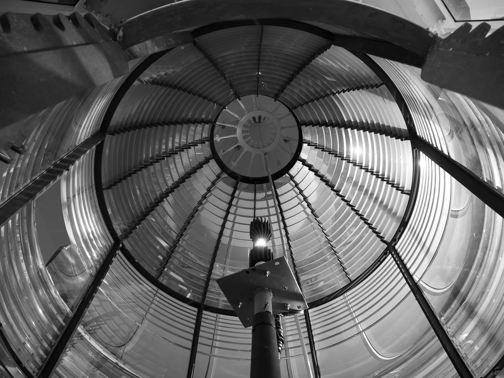 From the bottom perspective, at the top of a lighthouse. A spiral staircase with metal steps and railings encircles a central lamp, extending upwards within the cylindrical glass globe.
