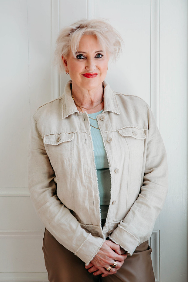 An older woman with short, messy white hair stands in front of a white paneled wall, smiling softly at the camera. She wears a light beige denim jacket over a mint green top and has rings on her fingers.