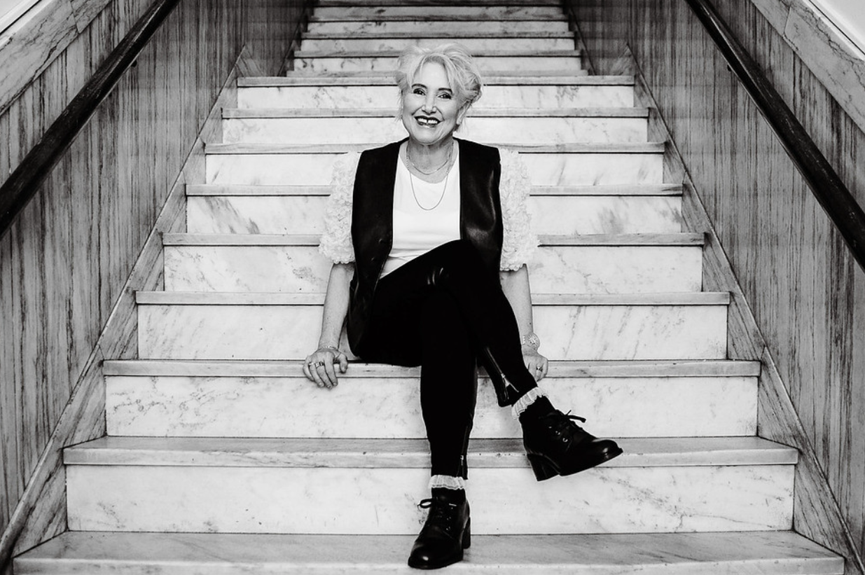 An elderly woman with short, styled hair sitting on a staircase made of marble, smiling at the camera. She wears a white top, black pants, a black vest, and black boots.