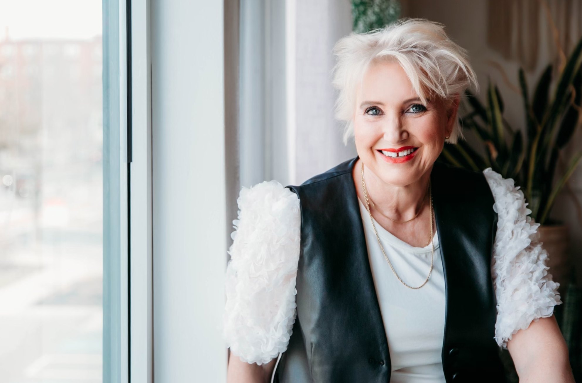 A smiling woman with short blonde hair and red lipstick, wearing a white top with textured sleeves and a black leather vest, standing indoors near a window with natural light.