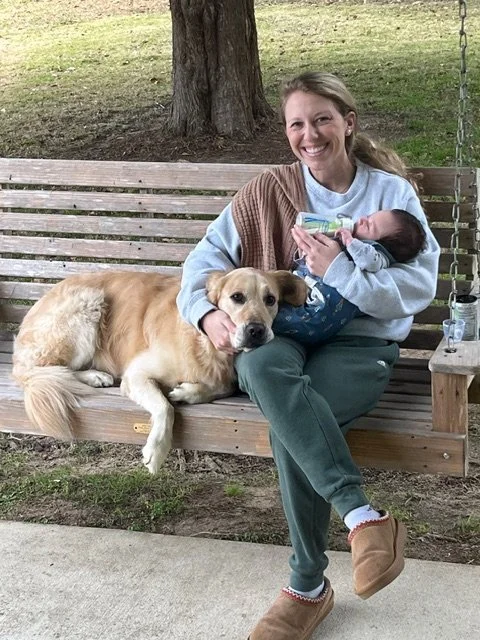 Woman sitting on a wooden bench holding a newborn baby and a dog lying beside her, outdoors near a tree.