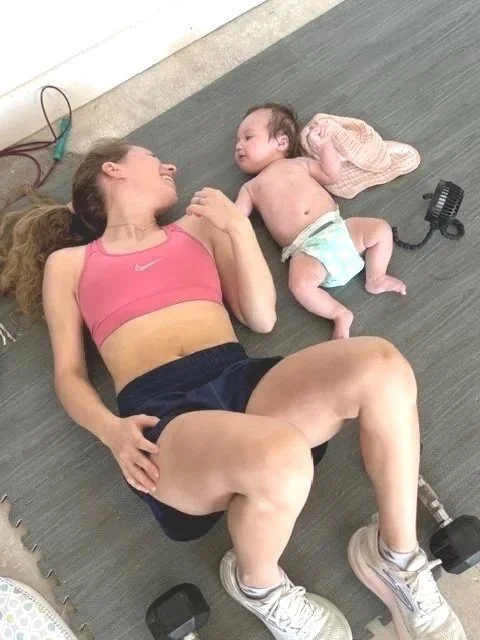 A woman and a baby lying on the floor, smiling and holding hands, with fitness equipment nearby.