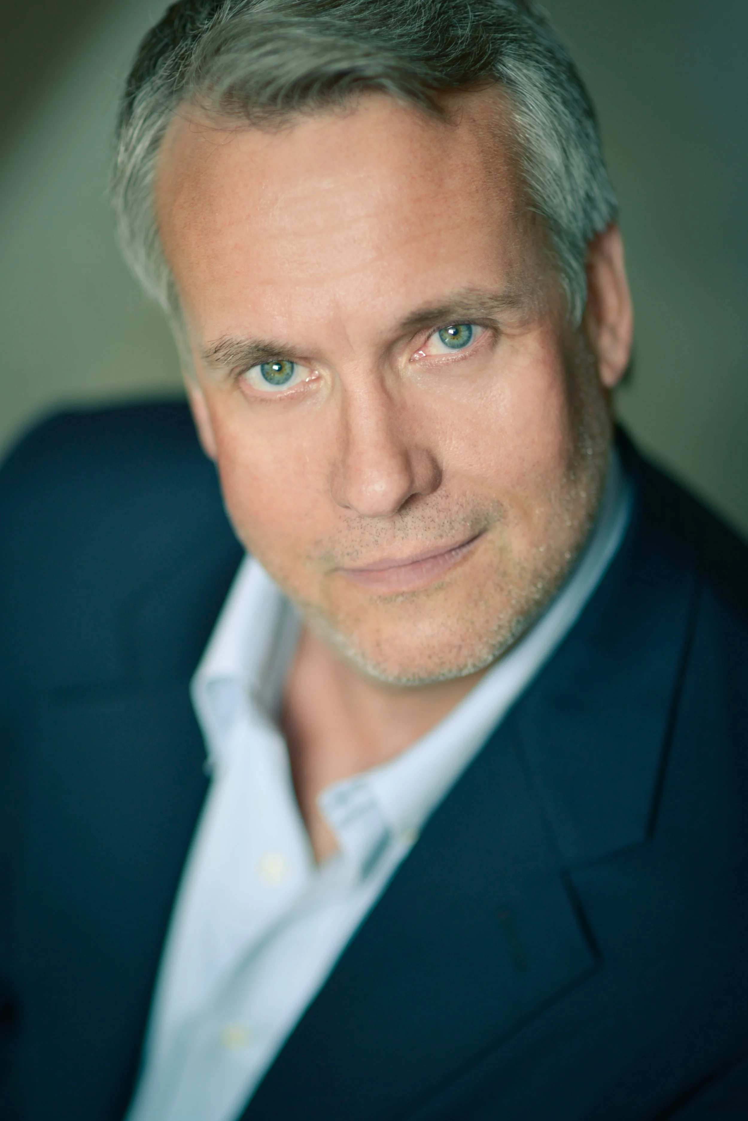Close-up portrait of a middle-aged man with short gray hair, blue eyes, wearing a navy blazer and white shirt, looking at the camera with a slight smile, against a blurred neutral background.