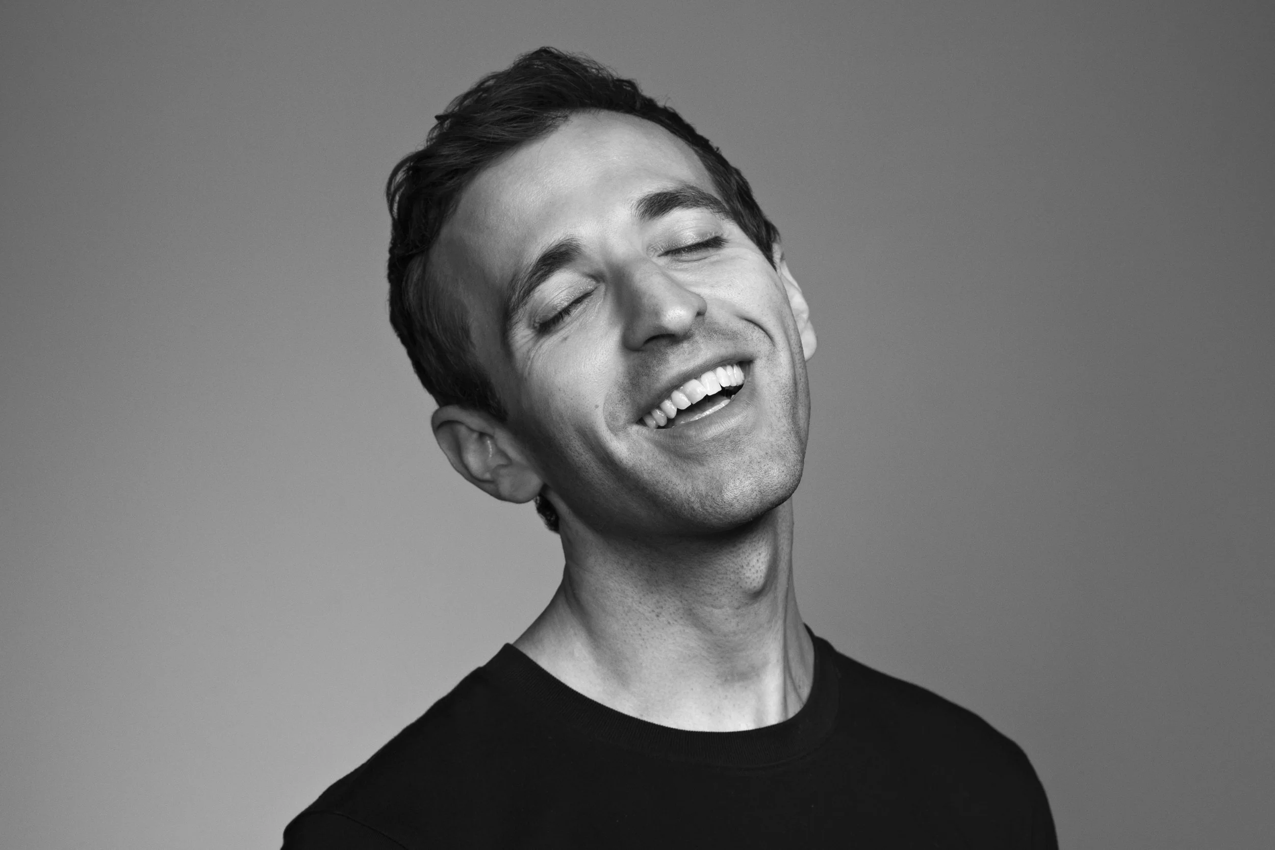Black and white photo of a young man laughing with eyes closed, wearing a black shirt, with a gradient background.
