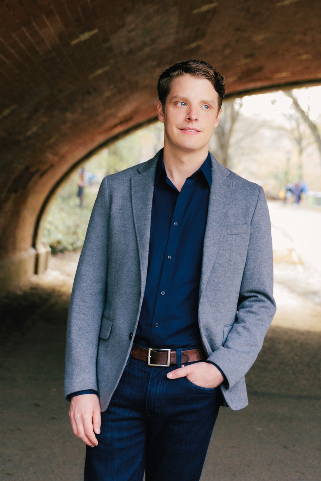 A young man with light skin and brown hair standing under a brick archway outdoors, dressed in a gray blazer, dark blue shirt, and jeans, with one hand in his pocket and looking to the side.