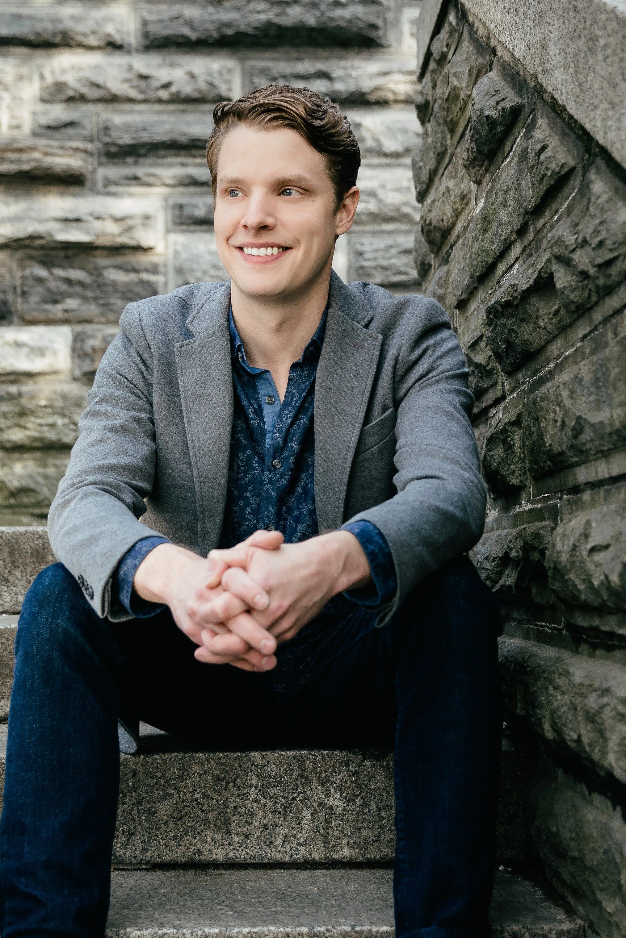 A young man with brown hair sits on steps, wearing a gray blazer and a blue shirt, smiling and looking to his left.