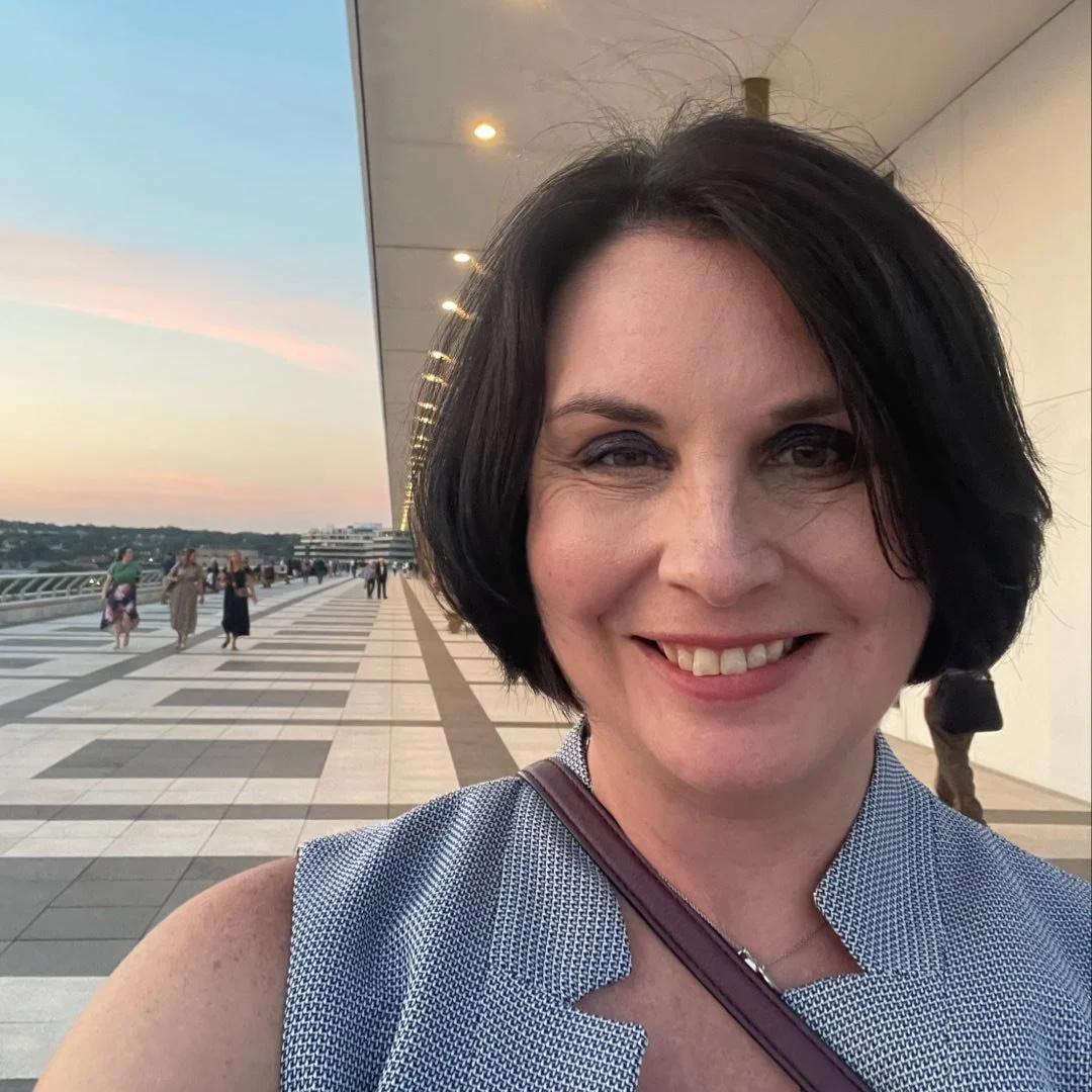 A smiling woman with short dark hair taking a selfie outdoors during sunset.