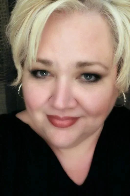 Close-up photo of a smiling woman with short blonde hair, blue eyes, and wearing dark makeup and a black top.