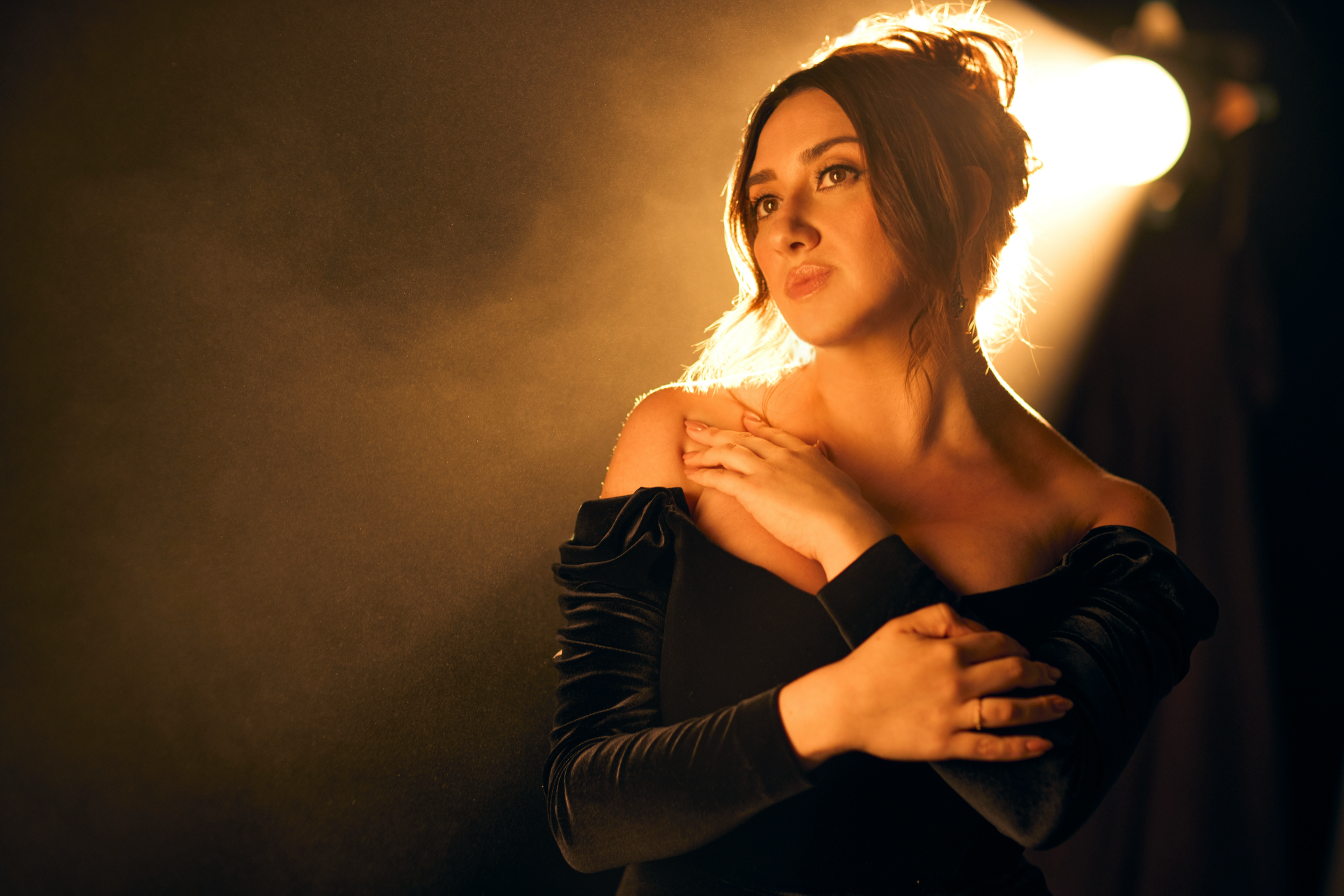 A woman with short, wavy brown hair is posing with her arms crossed over her shoulders, wearing a black off-the-shoulder dress, in front of a warm, backlit background.
