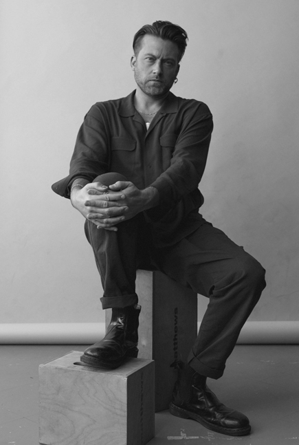 A man with a beard and styled hair sitting on a wooden box, holding a ball with both hands, wearing dark clothing and black boots, against a plain light background.