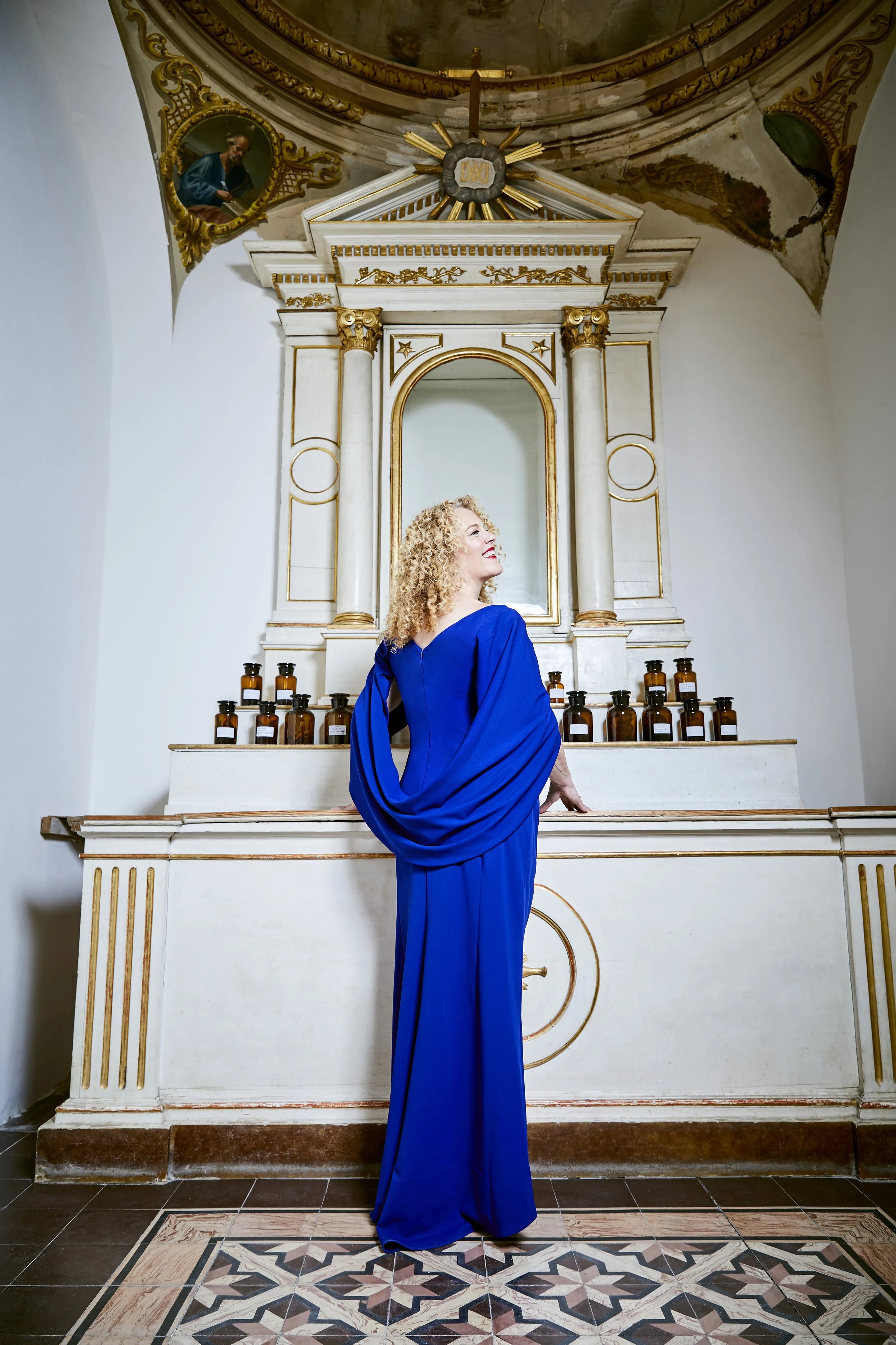 A woman in a bright blue dress standing in front of an ornate altar with multiple bottles on it inside a church.