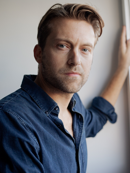 A young man with light skin and brown hair looking at the camera, wearing a denim shirt, standing indoors near a window.
