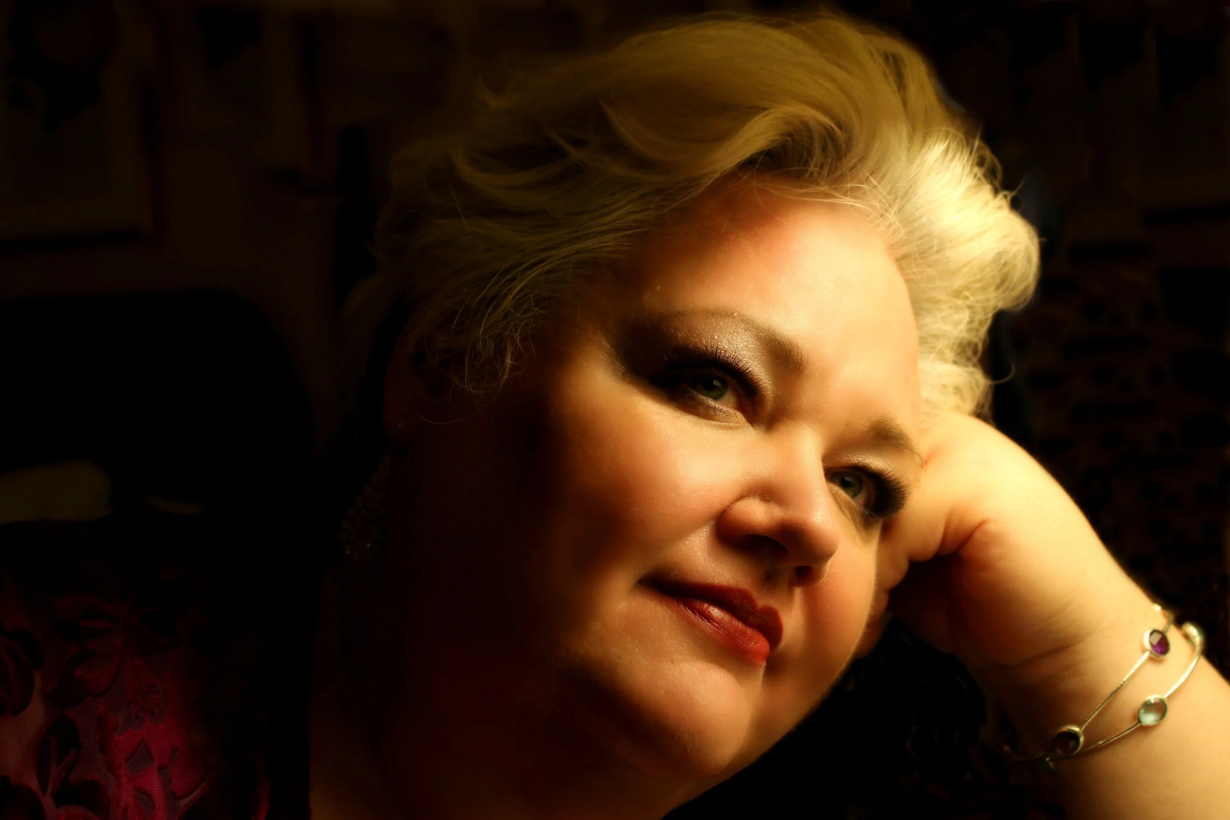 Close-up of a woman with blonde hair and makeup, resting her head on her hand in a dimly lit setting.