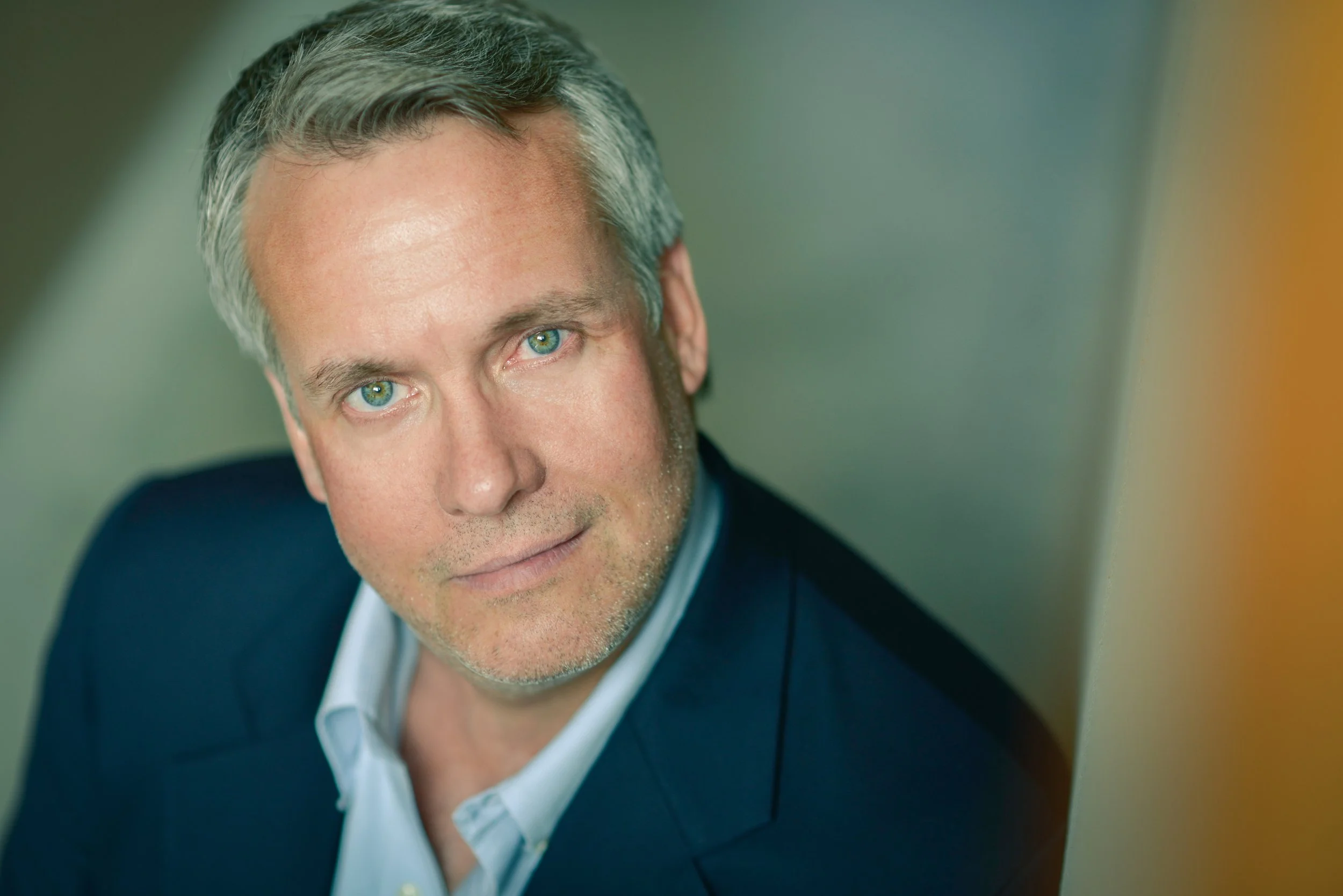 A middle-aged man with blue eyes, gray hair, and light stubble, wearing a dark suit and white shirt, looking at the camera with a slight smile.