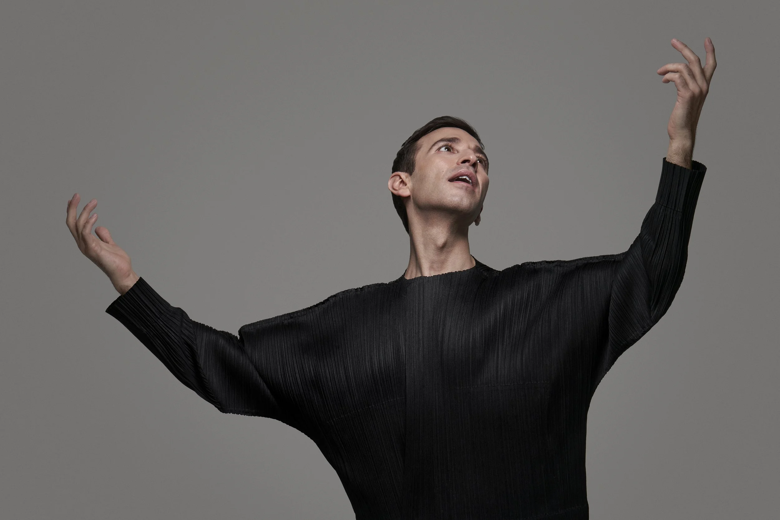 A man with dark hair, wearing a black pleated shirt, raises his arms and tilts his head back, looking upwards against a plain gray background.