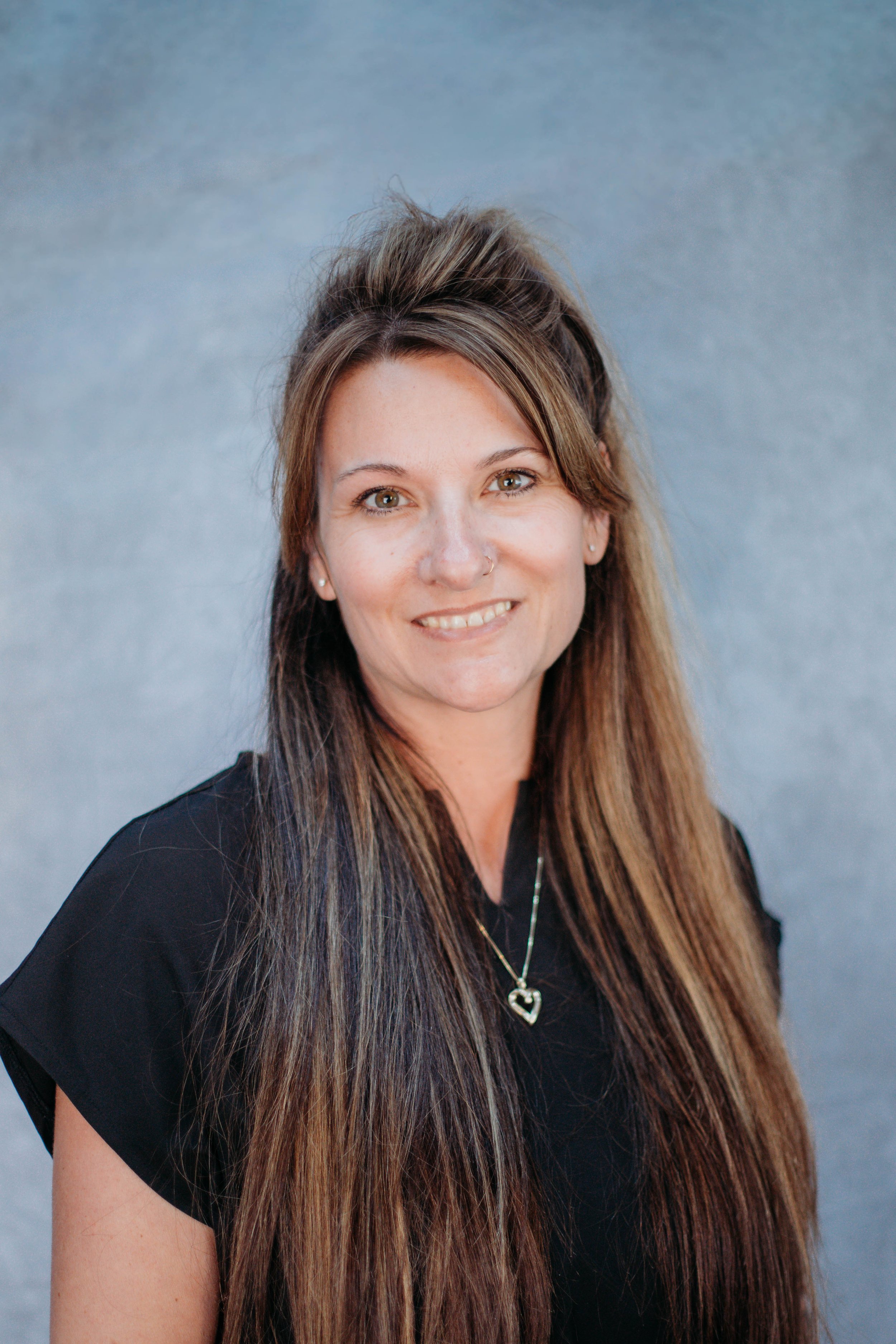 A woman with long brown hair, wearing a black shirt and a heart-shaped necklace, smiling against a gray background.