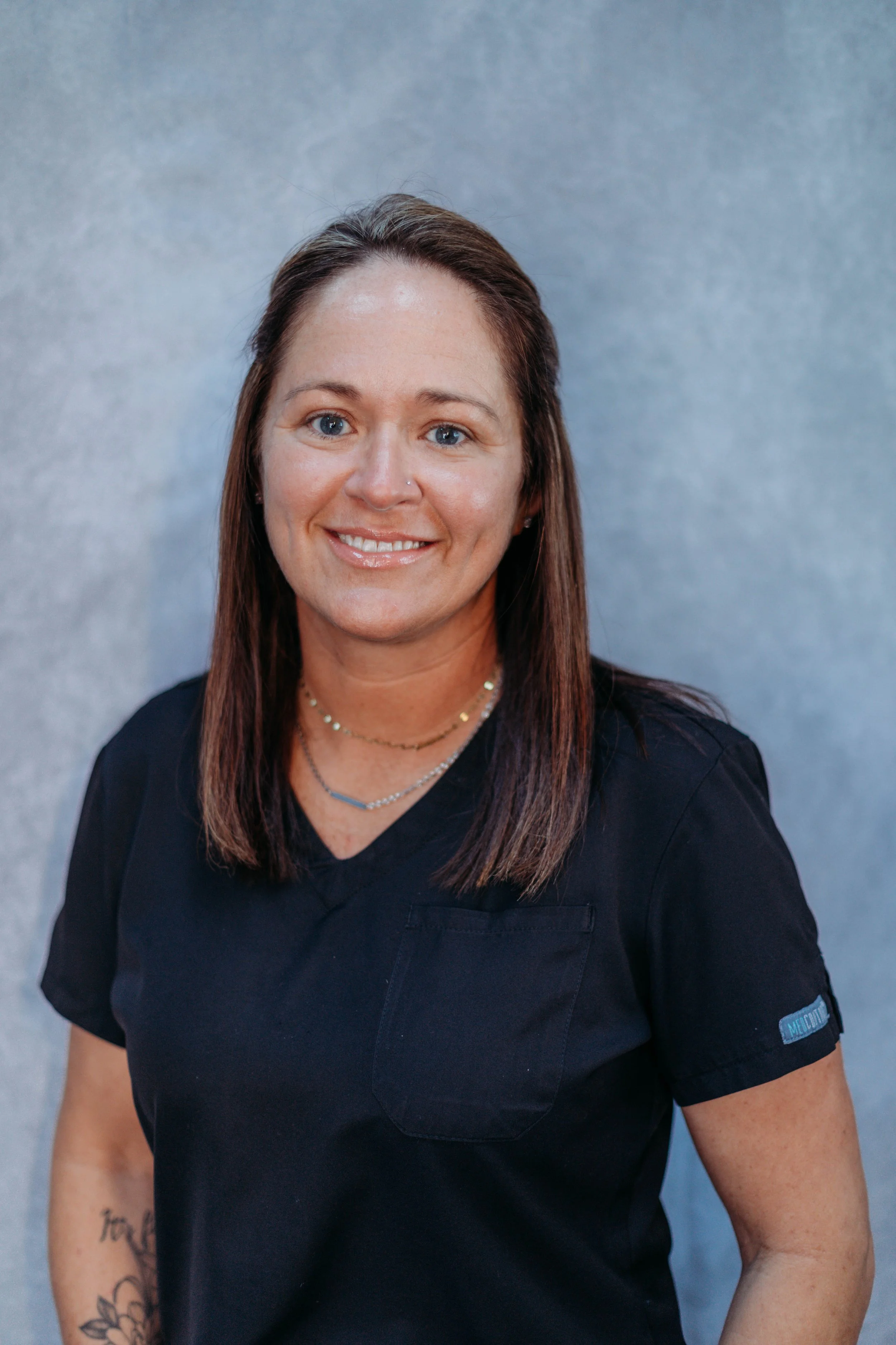 A woman with long brown hair wearing a black medical scrub top with a small logo on the sleeve, smiling at the camera against a gray background.