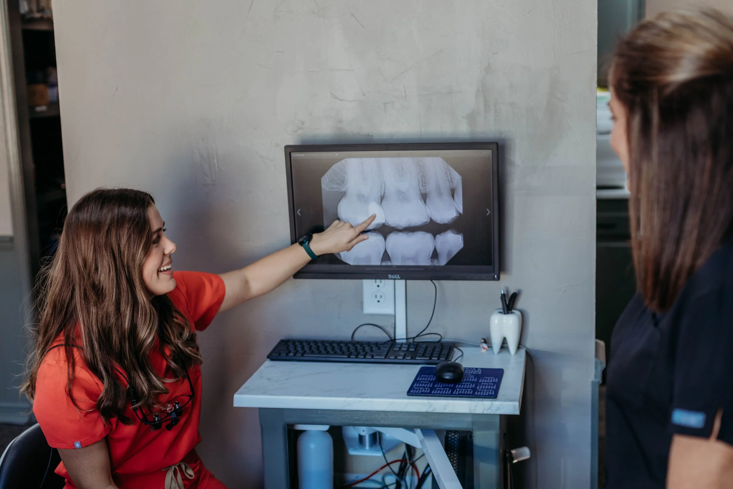 A woman in a red medical uniform showing a dental X-ray on a computer monitor to another person.