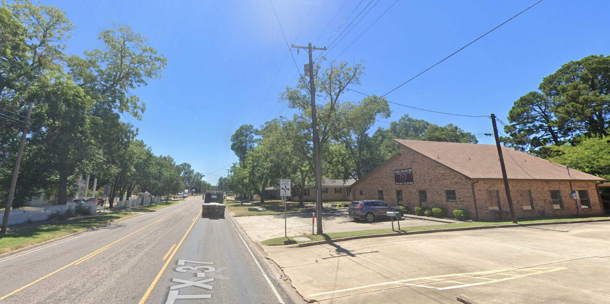 A sunny street scene with trees lining the road, a brick building on the right with a sign for a dental office, cars parked along the sidewalk, and a clear blue sky overhead.