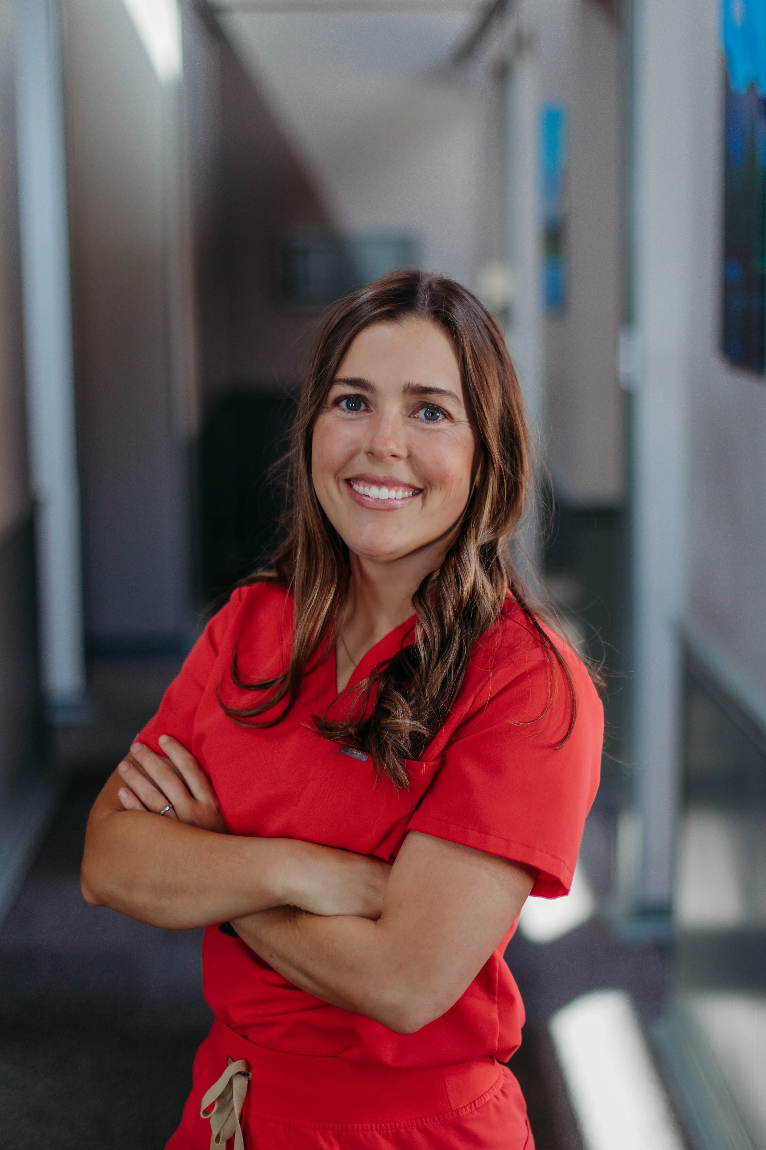 A smiling woman in an orange scrubs with her arms crossed, standing in a hallway with blue artwork on the walls.