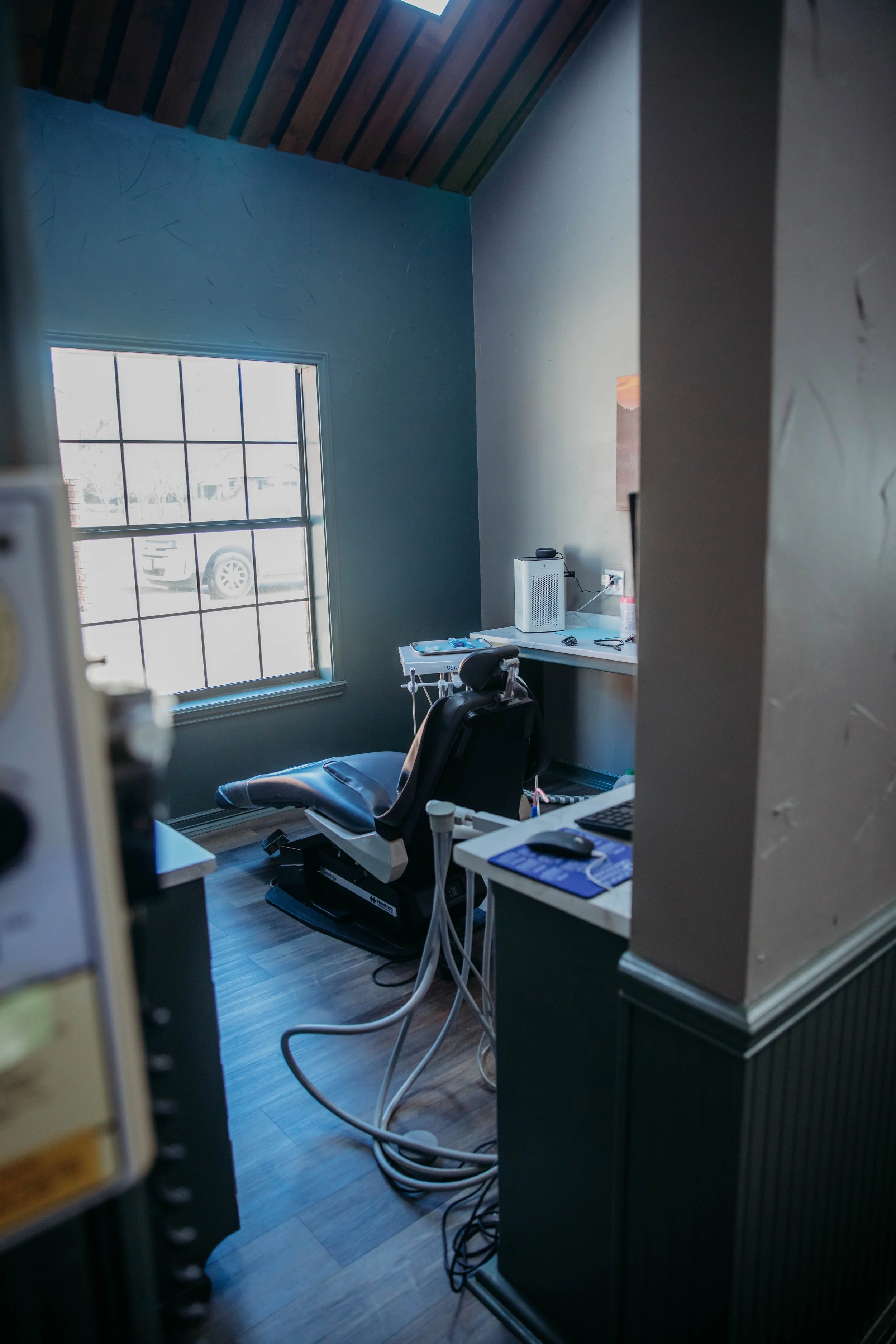 A medical examination room with a window, a black examination chair, and various medical dental  equipment on a table, including a monitor and wires.