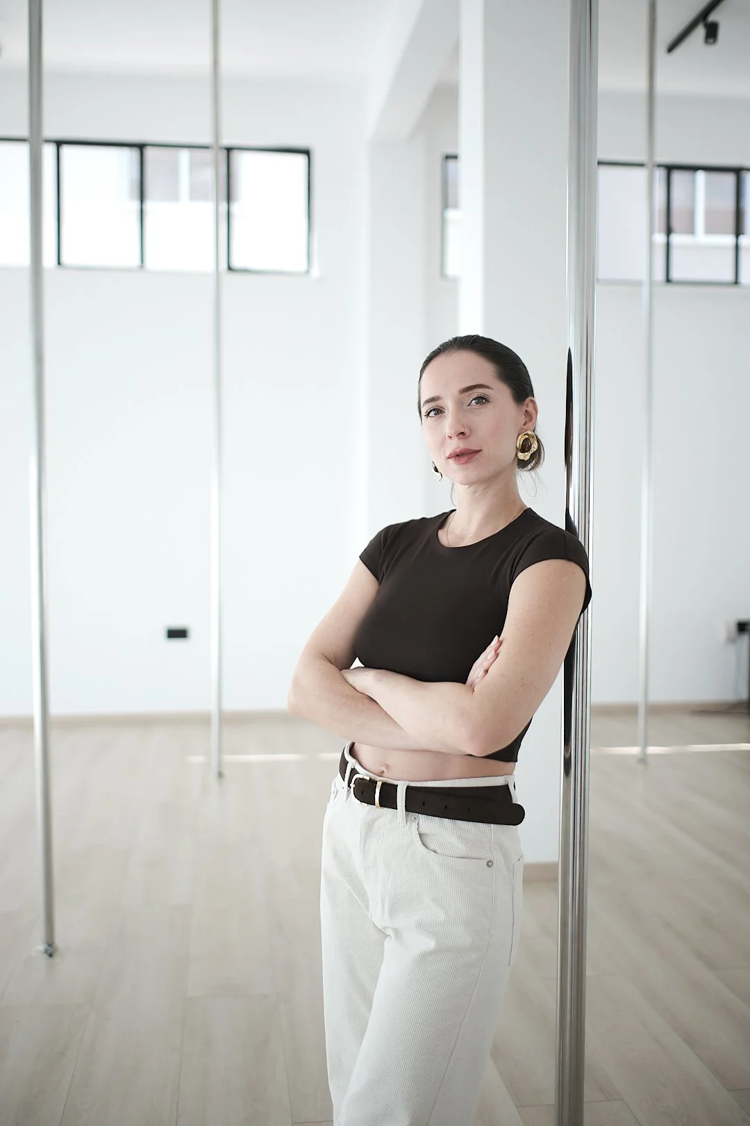 A young woman with dark hair, wearing a black crop top, white pants, and gold earrings, stands with crossed arms next to a mirror in a bright dance or fitness studio with large windows.