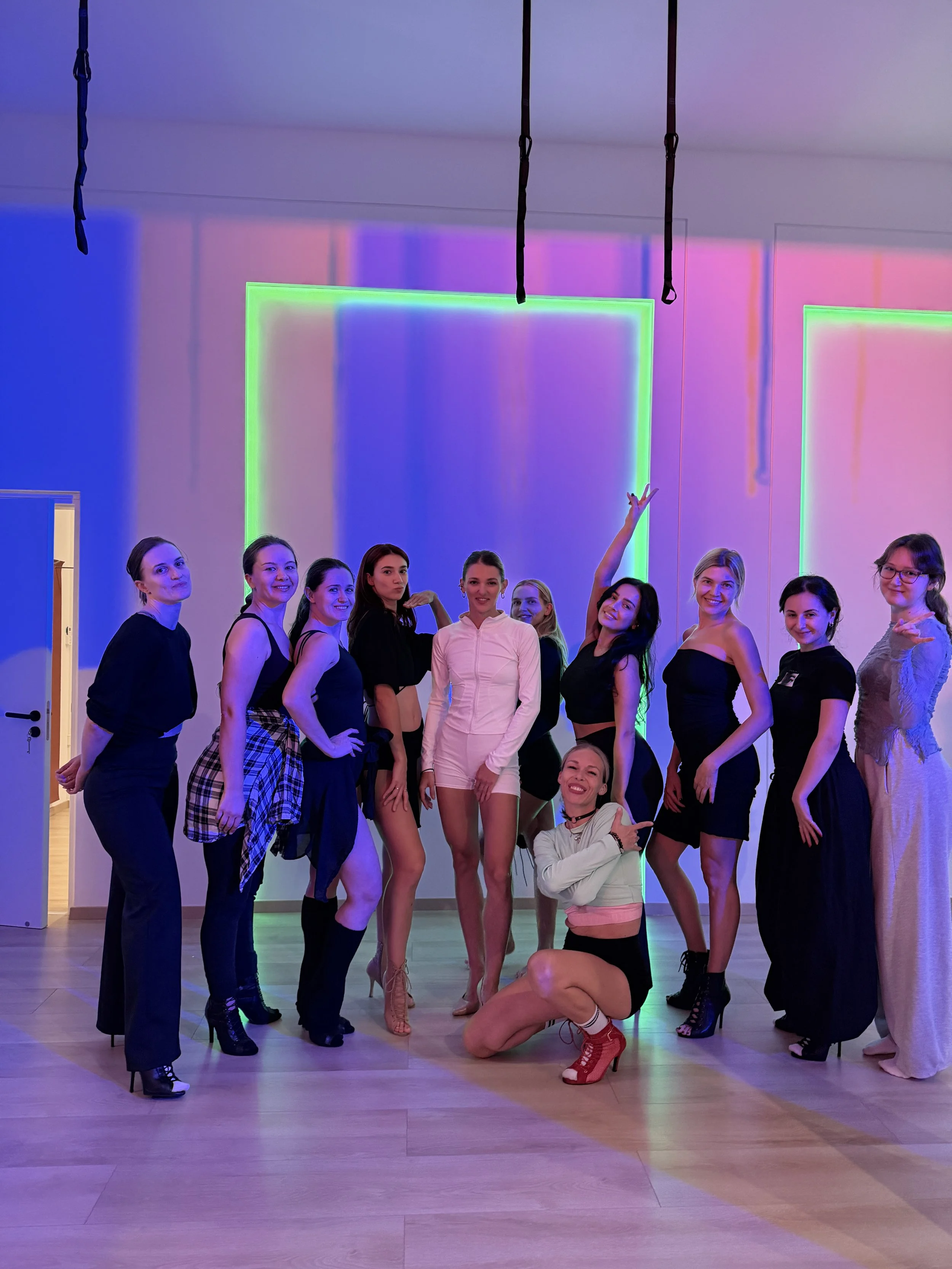 A group of women posing together in a dance studio with colorful neon lights on the wall, some standing and one kneeling in front, all smiling and looking at the camera.
