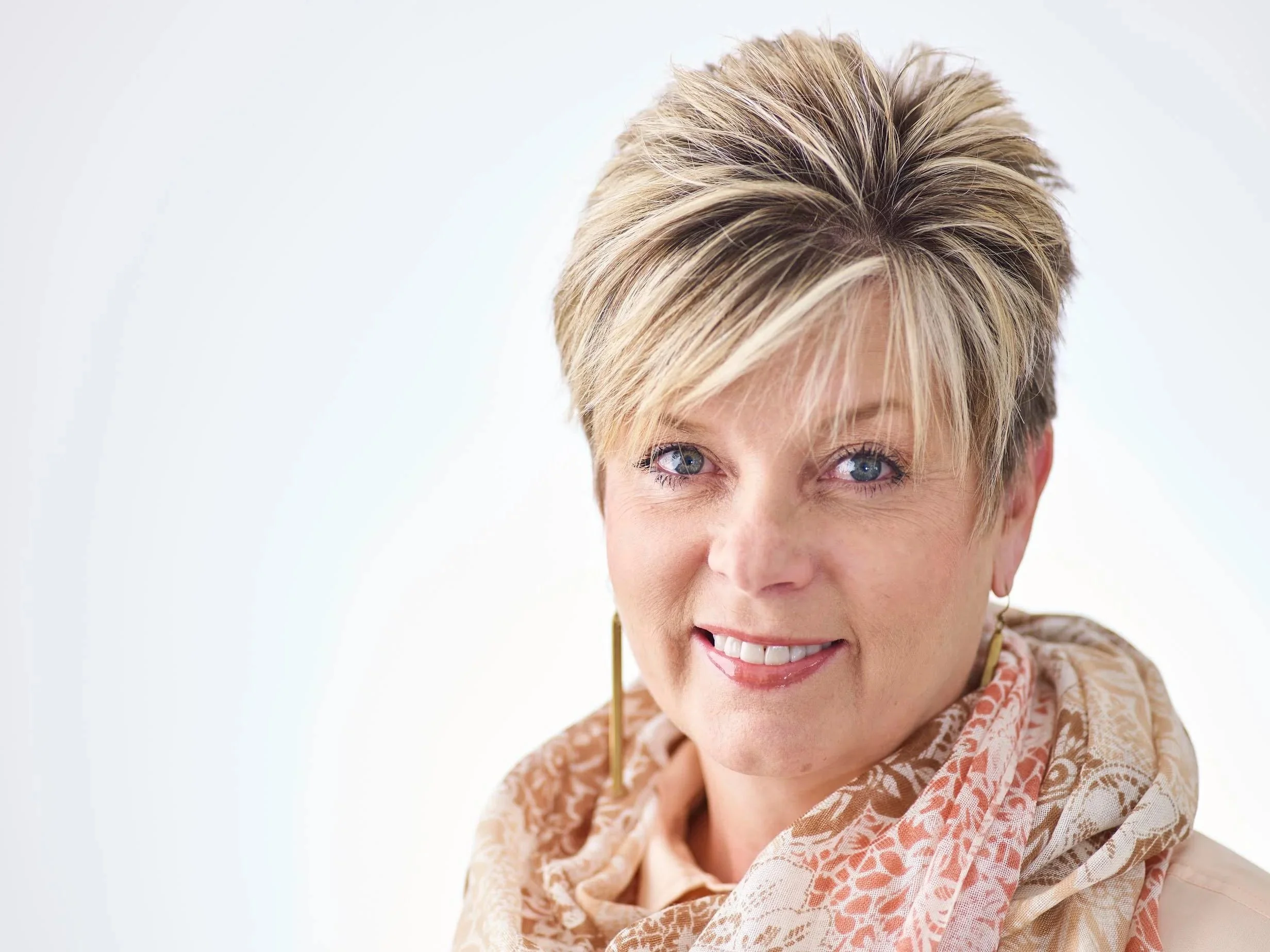 A woman with short blonde hair and blue eyes smiling at the camera, wearing a patterned beige shawl and gold earrings.