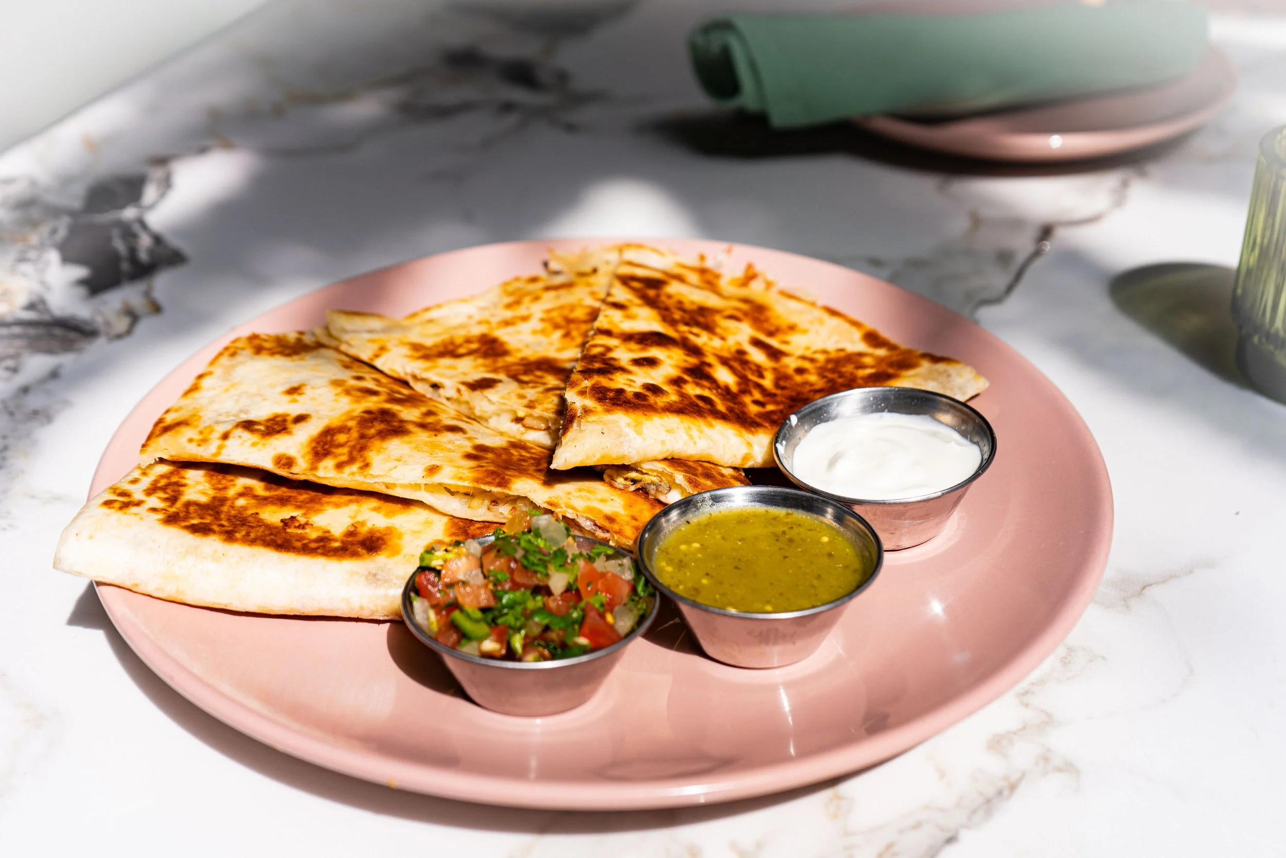 A pink plate with cheese quesadilla slices, topped with dark grill marks, accompanied by small bowls of salsa, green sauce, and sour cream, on a marble countertop.