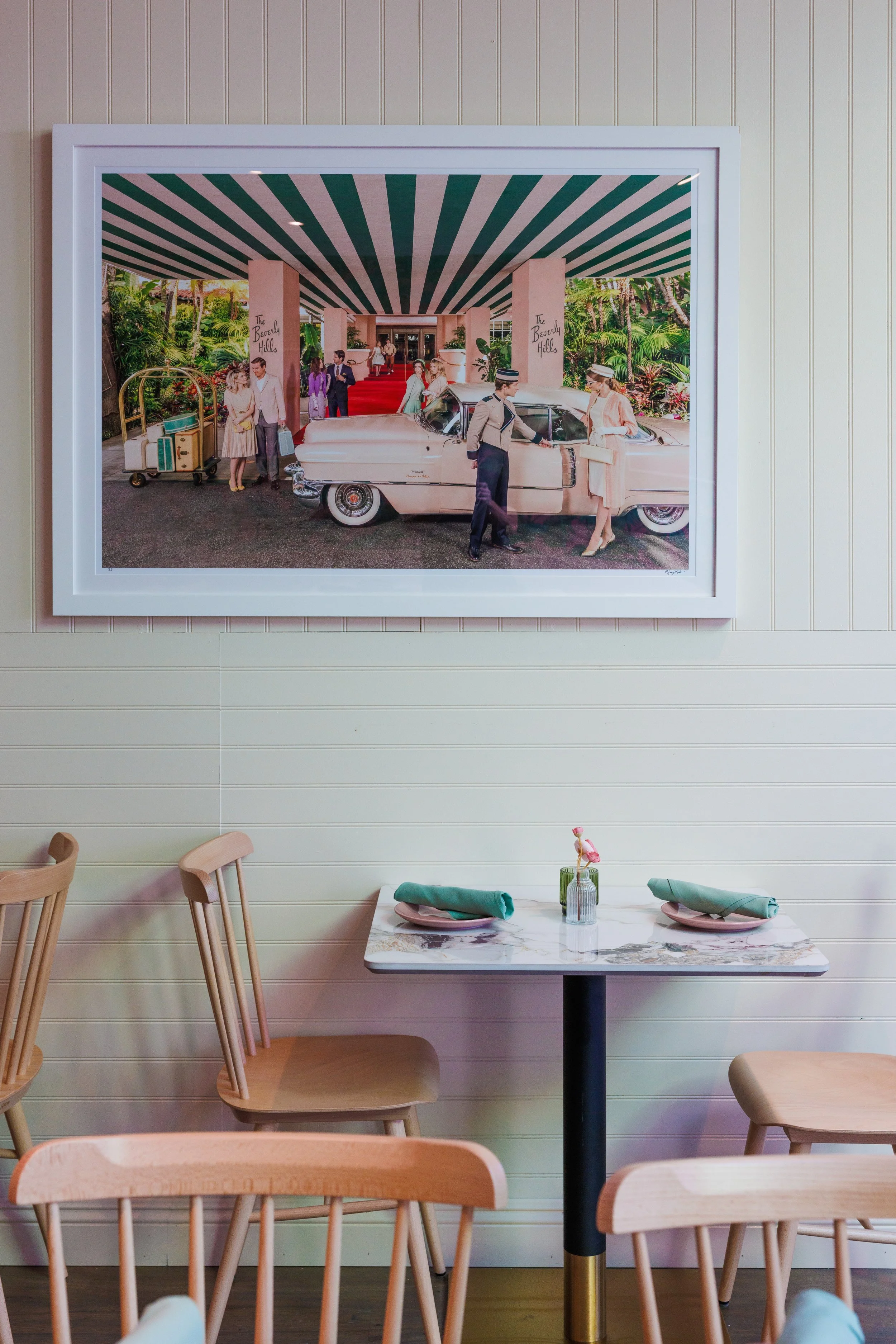 A framed vintage-style photograph of a hotel entrance with a couple greeting a doorman, alongside a Classic car, on a wall inside a restaurant with a small table set for two in the foreground.