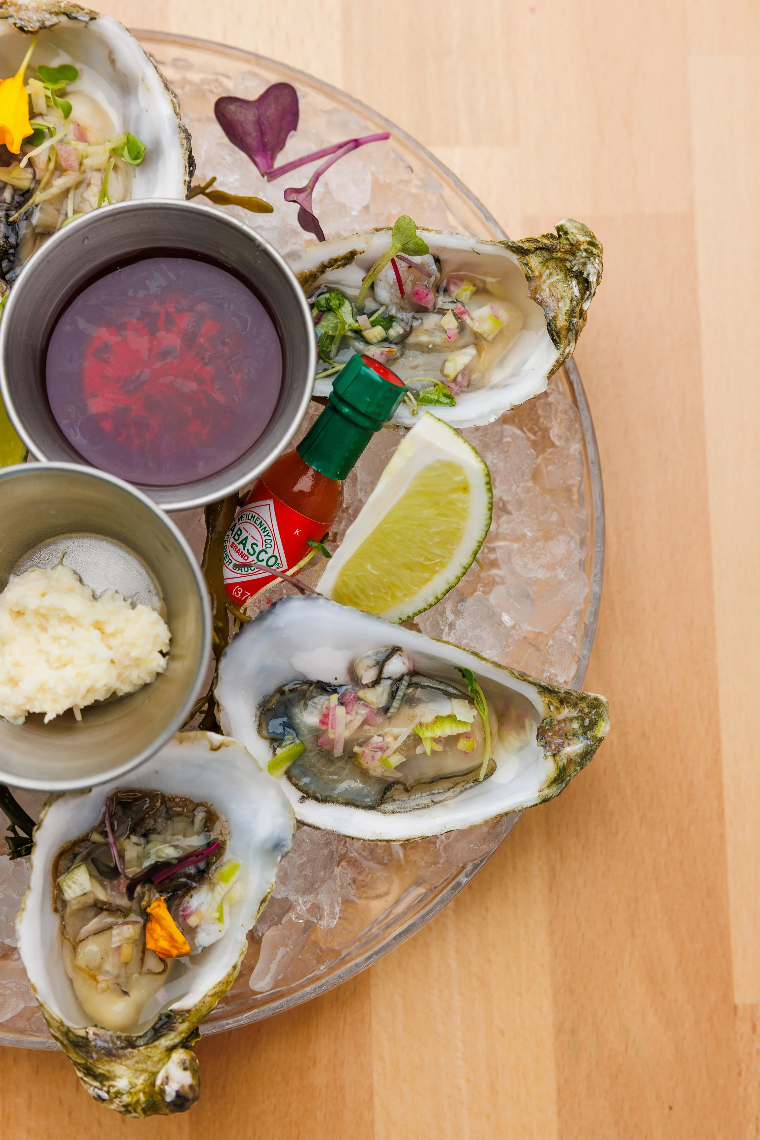 A plate of fresh oysters served on crushed ice, garnished with lemon wedges, microgreens, and various sauces including hot sauce and mignonette poured in small bowls.