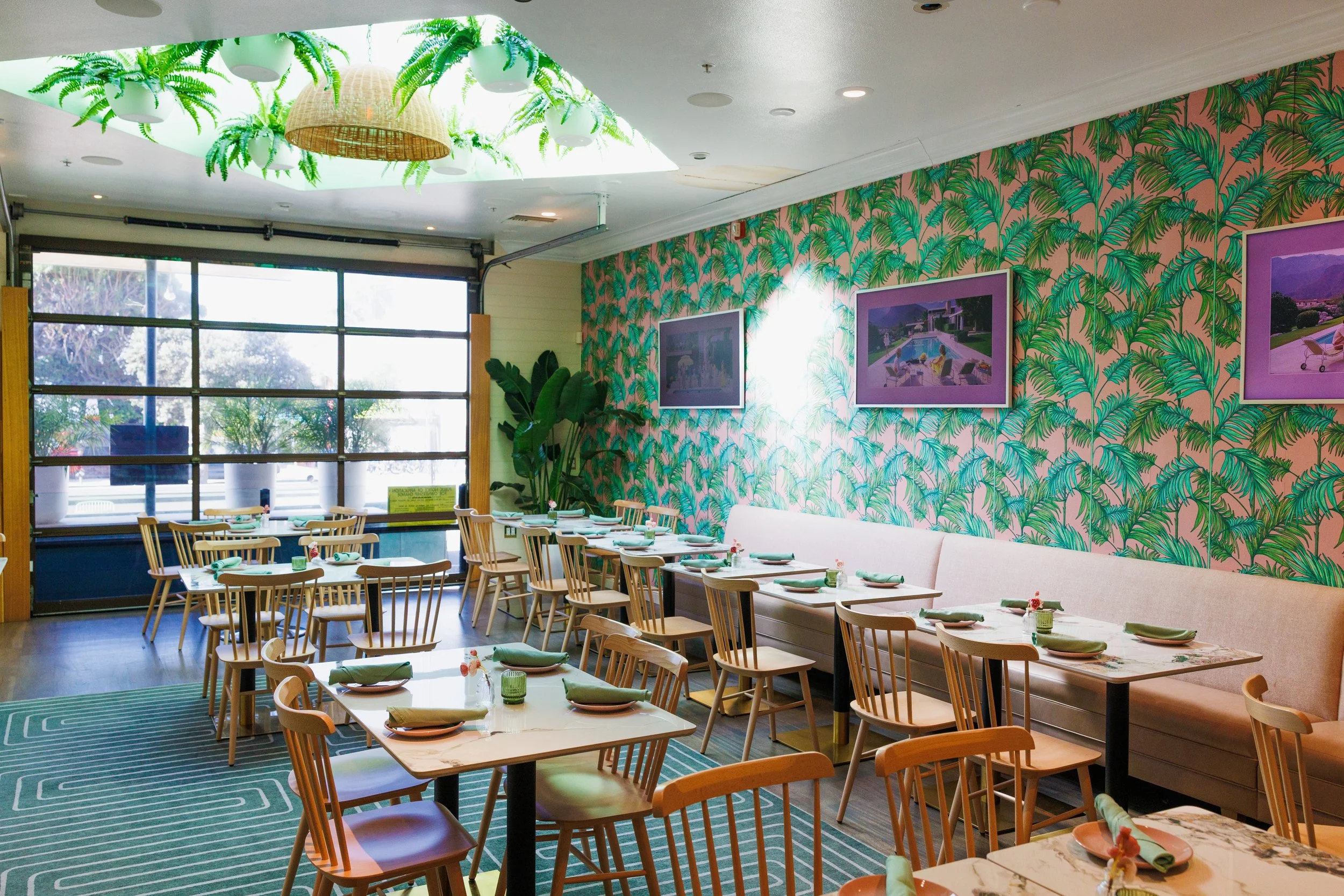 Interior of a restaurant with wooden chairs and tables set with plates, napkins, and small flower arrangements. The wall features pink and green tropical leaf patterned wallpaper, with framed pictures hanging. A large window lets in natural light, and a ceiling decoration mimics green tropical plants.